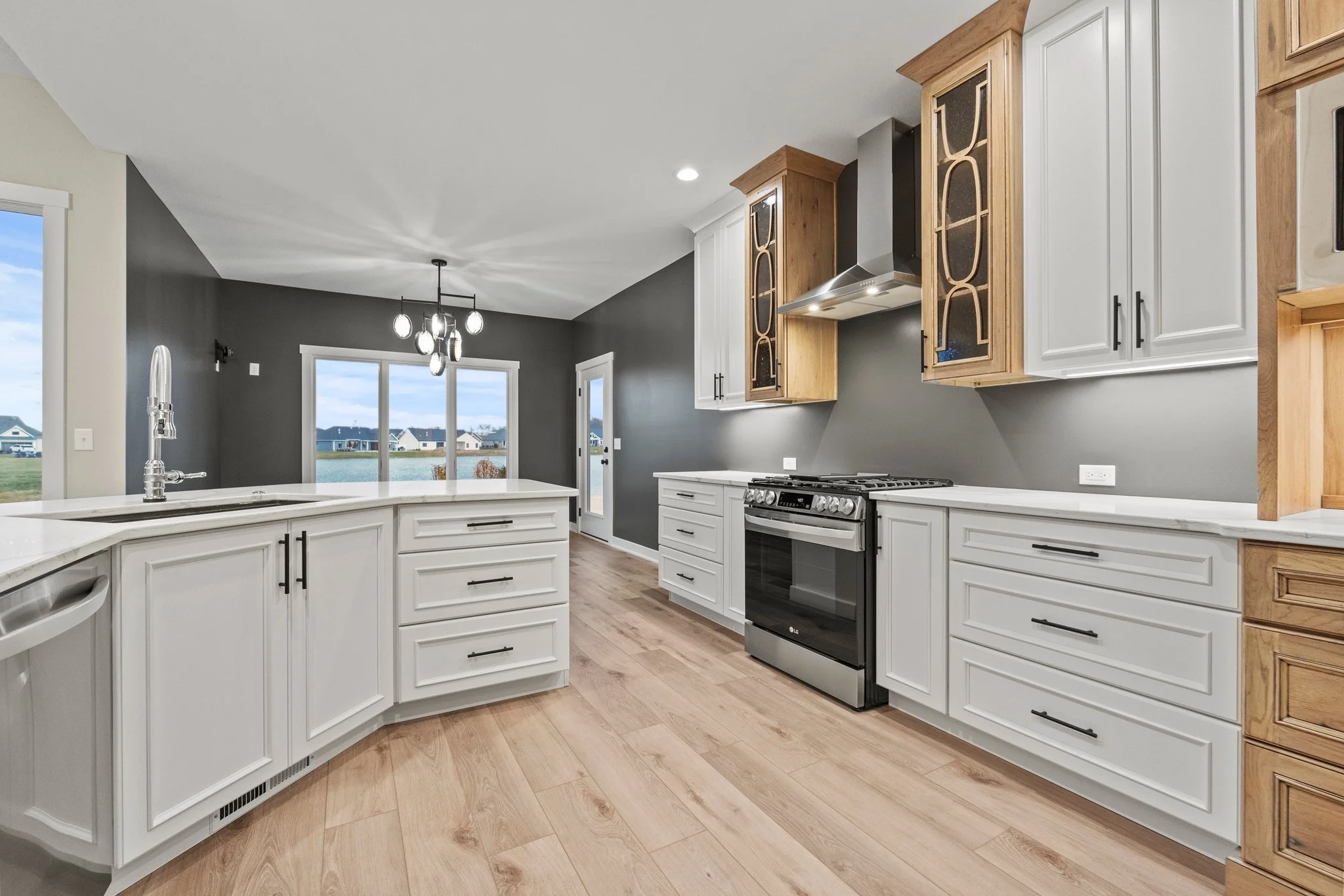 Modern kitchen with white cabinets, stainless steel stove, wood accents, gray walls, and hardwood floors, overlooking a dining area with large windows and a glass door to an outdoor view.