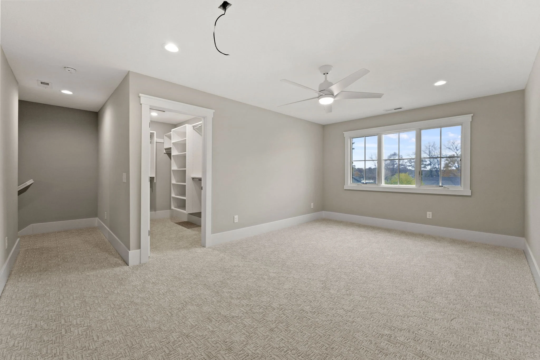 Empty living room with beige carpet, white baseboards, beige walls, large window showing trees outside, ceiling fan with light, and closet with shelving and hanging space.