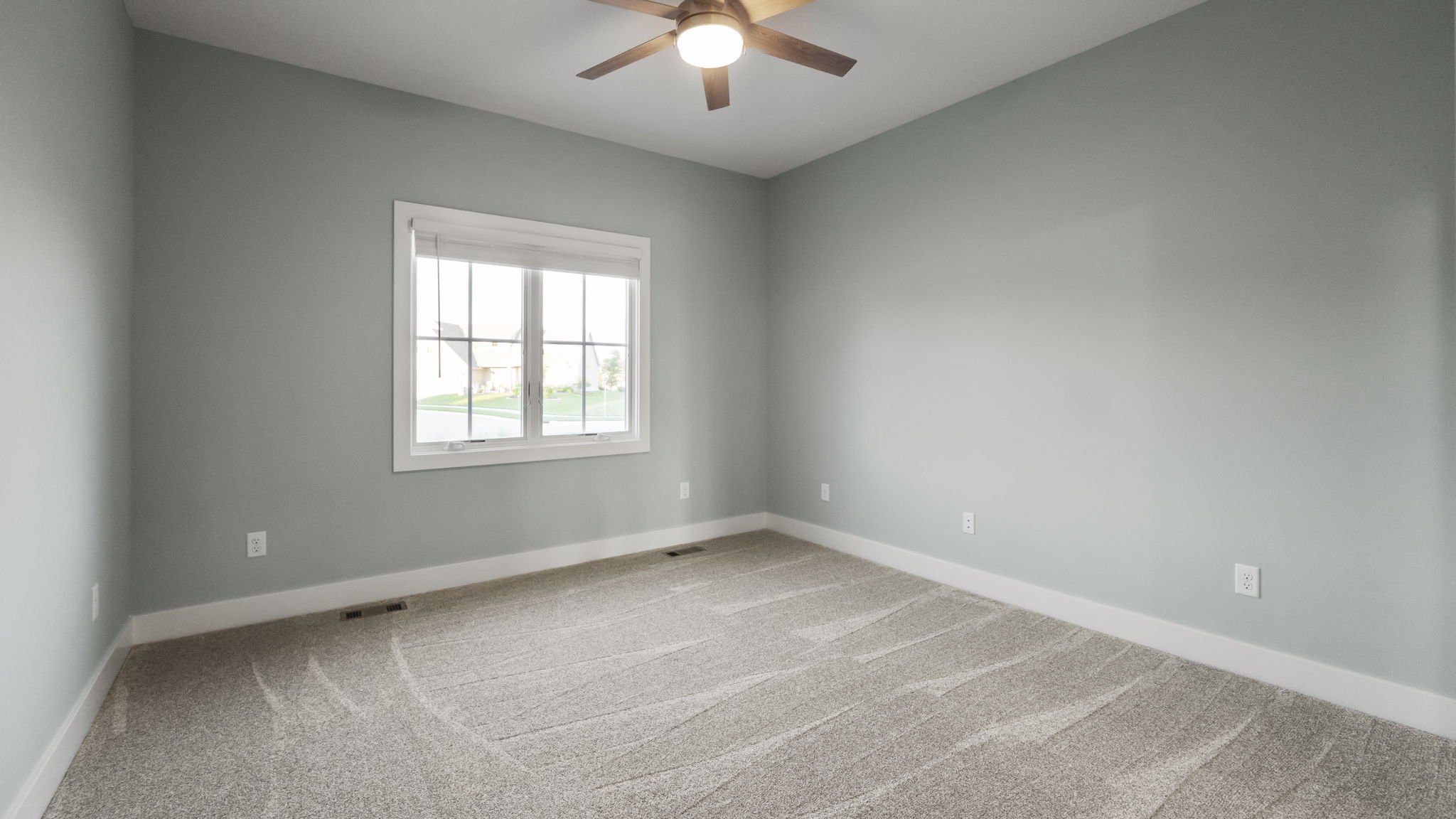 Empty bedroom with light green walls, beige carpet, a window showing a suburban neighborhood, a ceiling fan, and multiple electrical outlets.