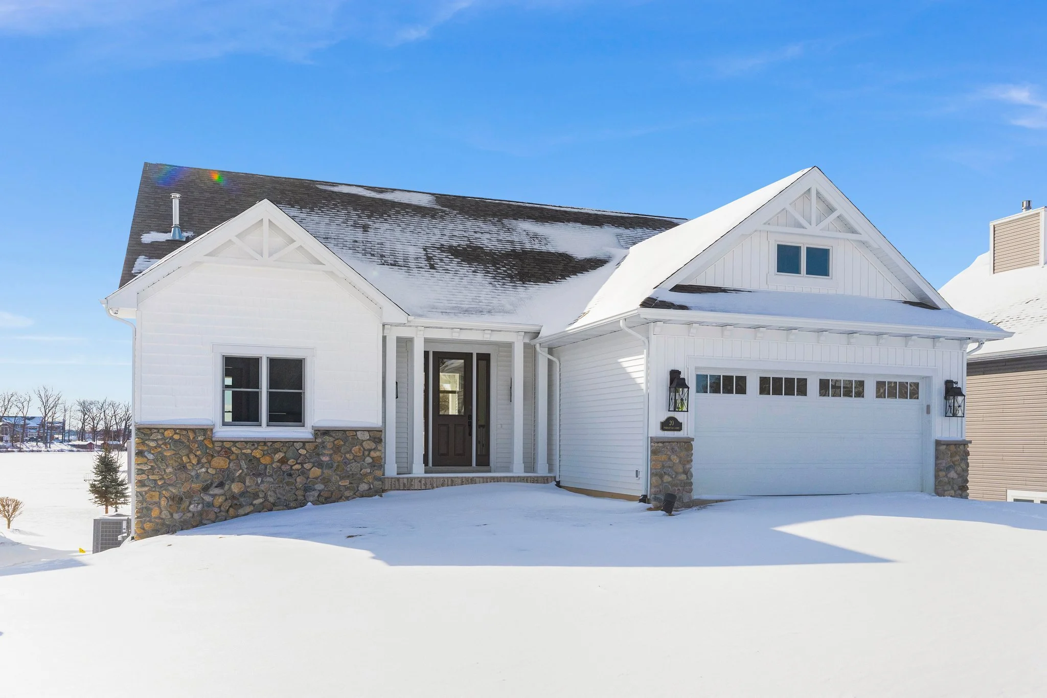 White house with stone accents on lower walls, black front door, attached garage, snow-covered lawn, and a clear blue sky.
