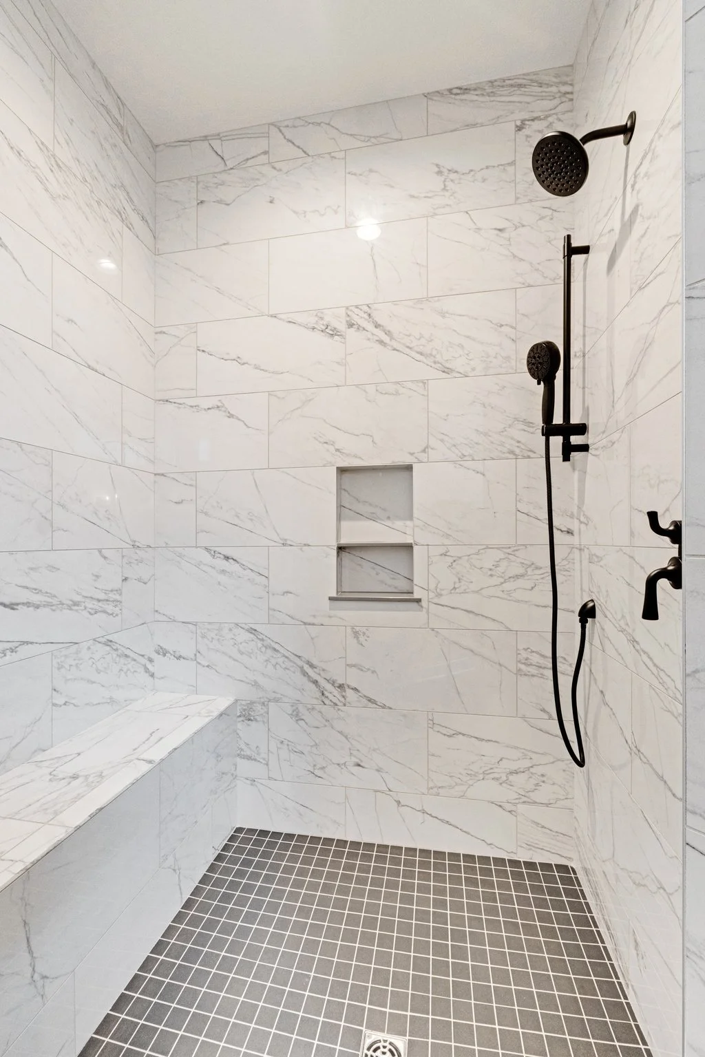 A modern walk-in shower with white marble walls, black fixtures, a built-in bench, a recessed shelf, and a black showerhead with a hand-held spray, beneath a ceiling light.