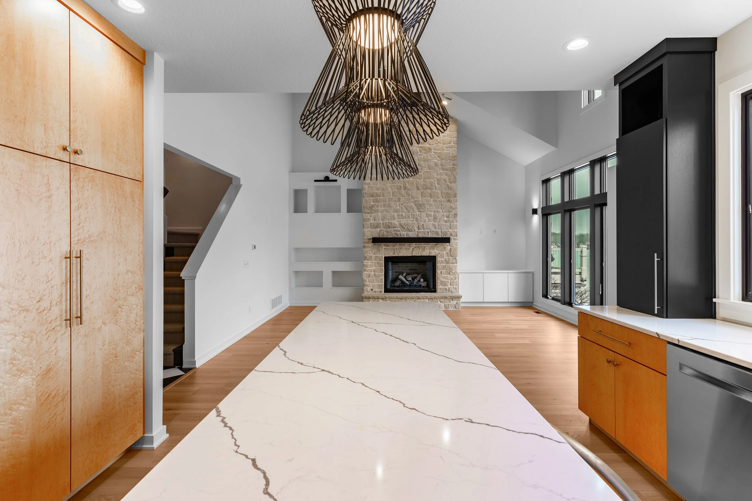 Modern living room with a stone fireplace, large black and wooden cabinetry, hardwood floors, and large windows with black framing.