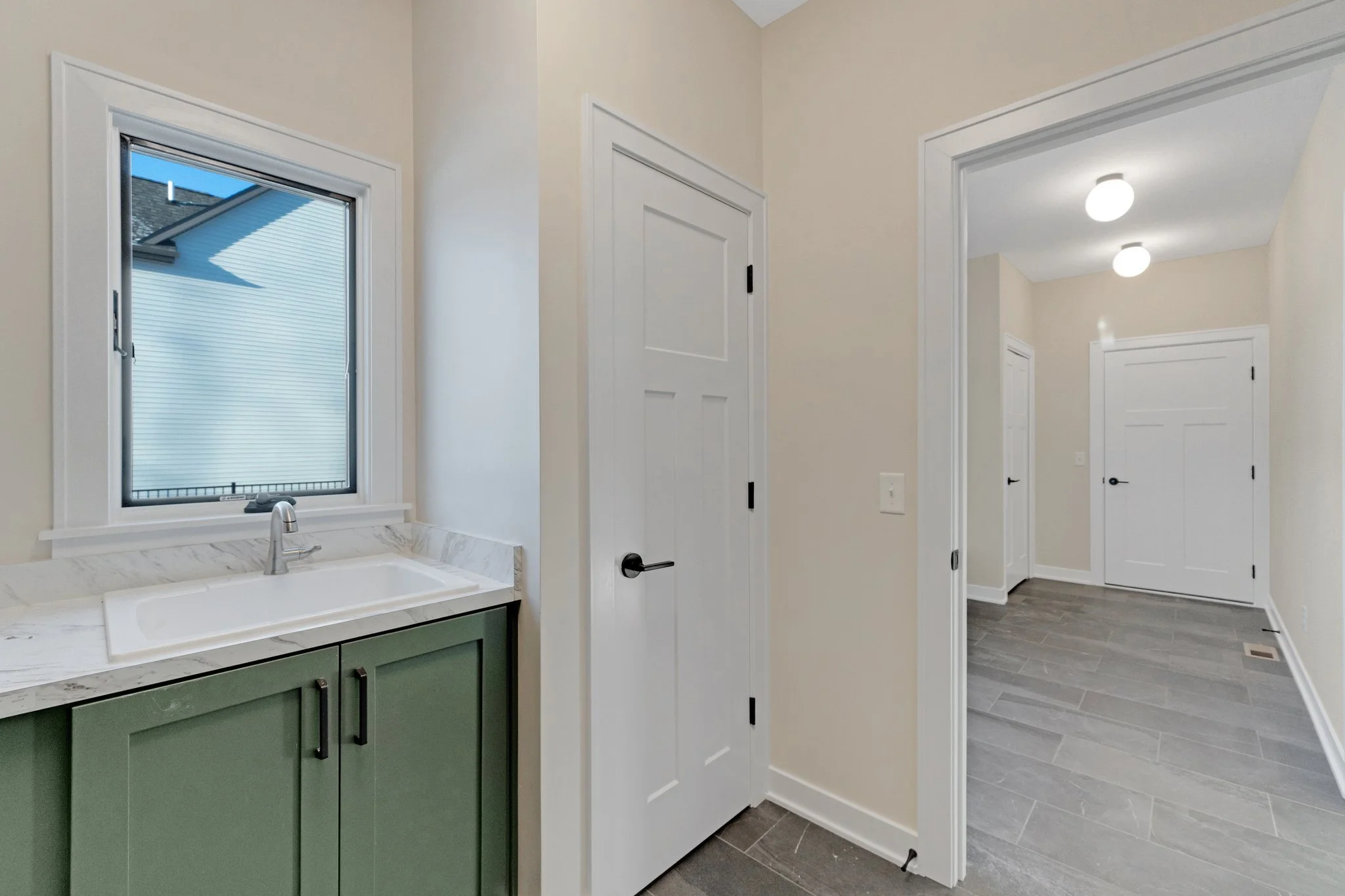 Interior view of a house with a small laundry room or utility area that has a green cabinet with a marble countertop and a white sink, a window with a blind showing part of the exterior, beige walls, white doors, and gray wood-like flooring.