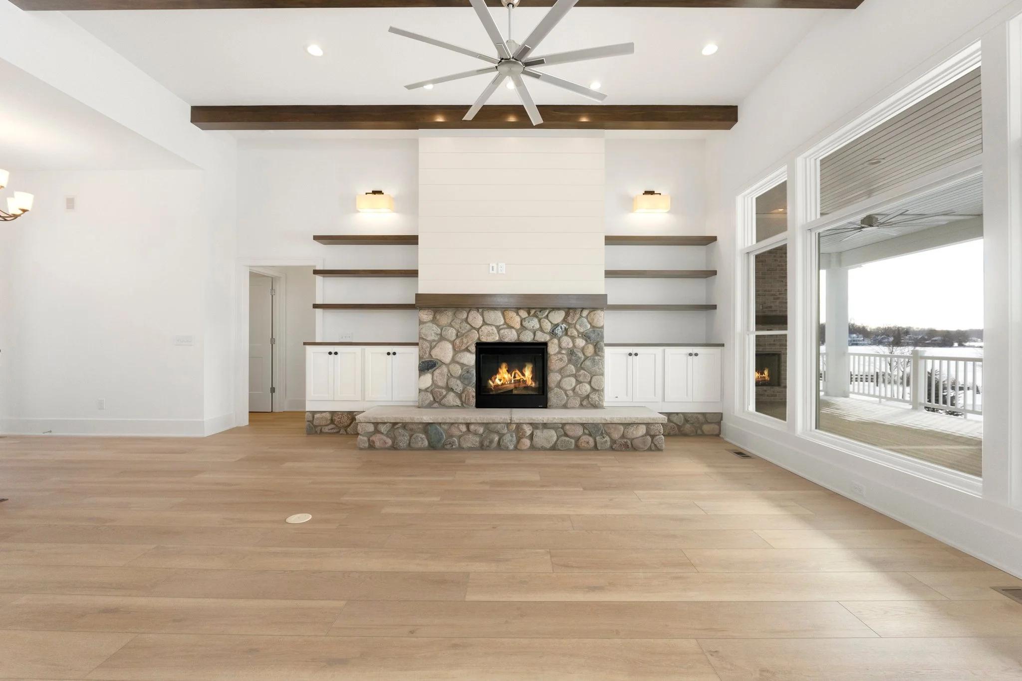 Empty living room with a stone fireplace, wooden shelves, large windows, and a ceiling fan.