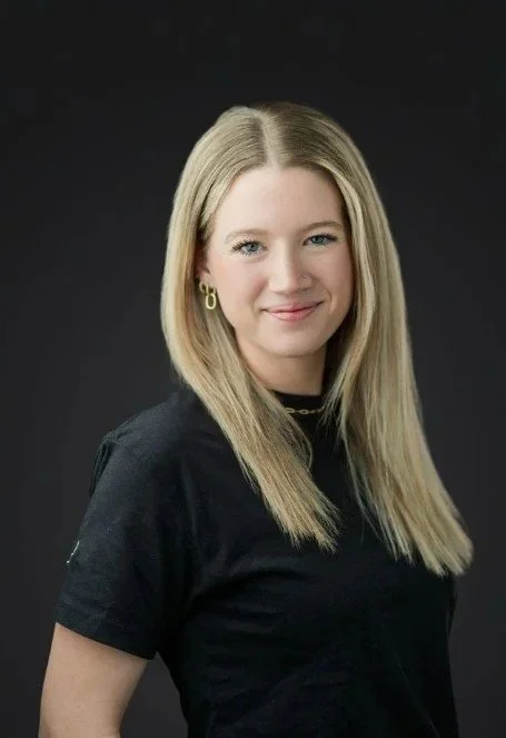 A young woman with blonde hair, wearing a black shirt and gold hoop earrings, smiling at the camera against a dark background.