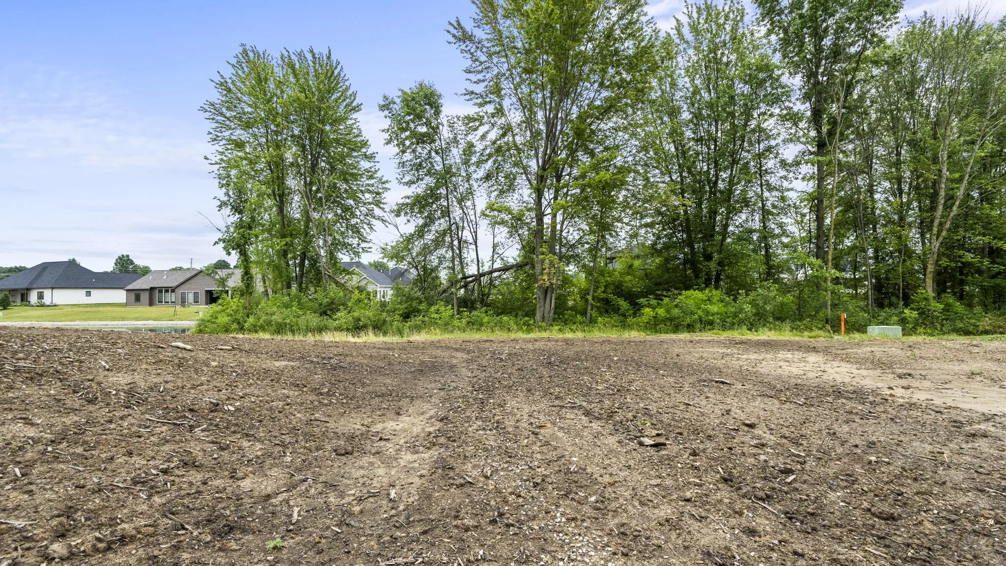 A cleared dirt lot in the foreground with a line of trees and green foliage behind, with residential houses in the background under a partly cloudy sky.