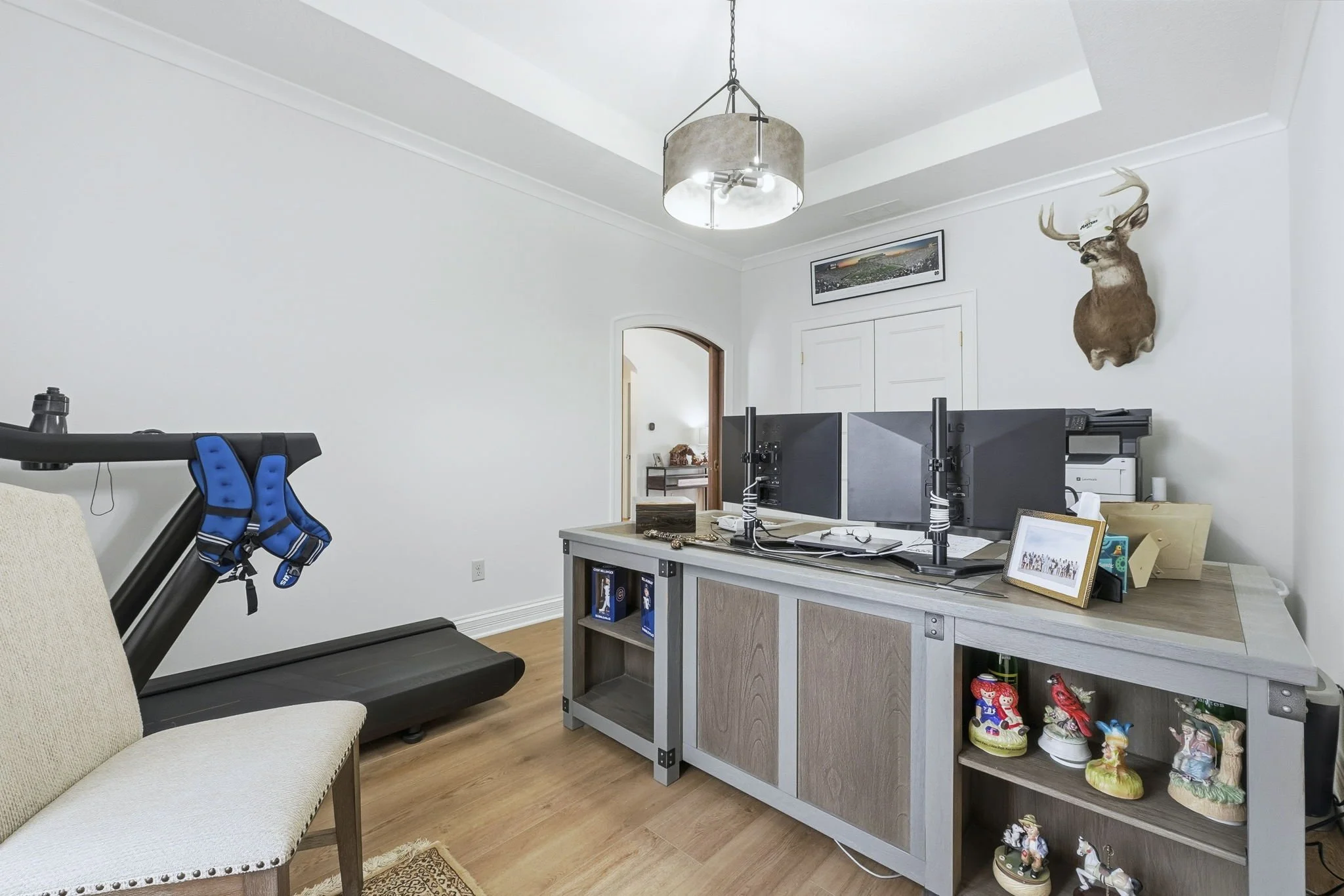 Home office with a treadmill on the left, a wooden desk with three computer monitors, a framed photo, and decorative items, and a wall-mounted deer head with a cap on it. There is a ceiling light fixture, a white door, and a hallway visible in the ba