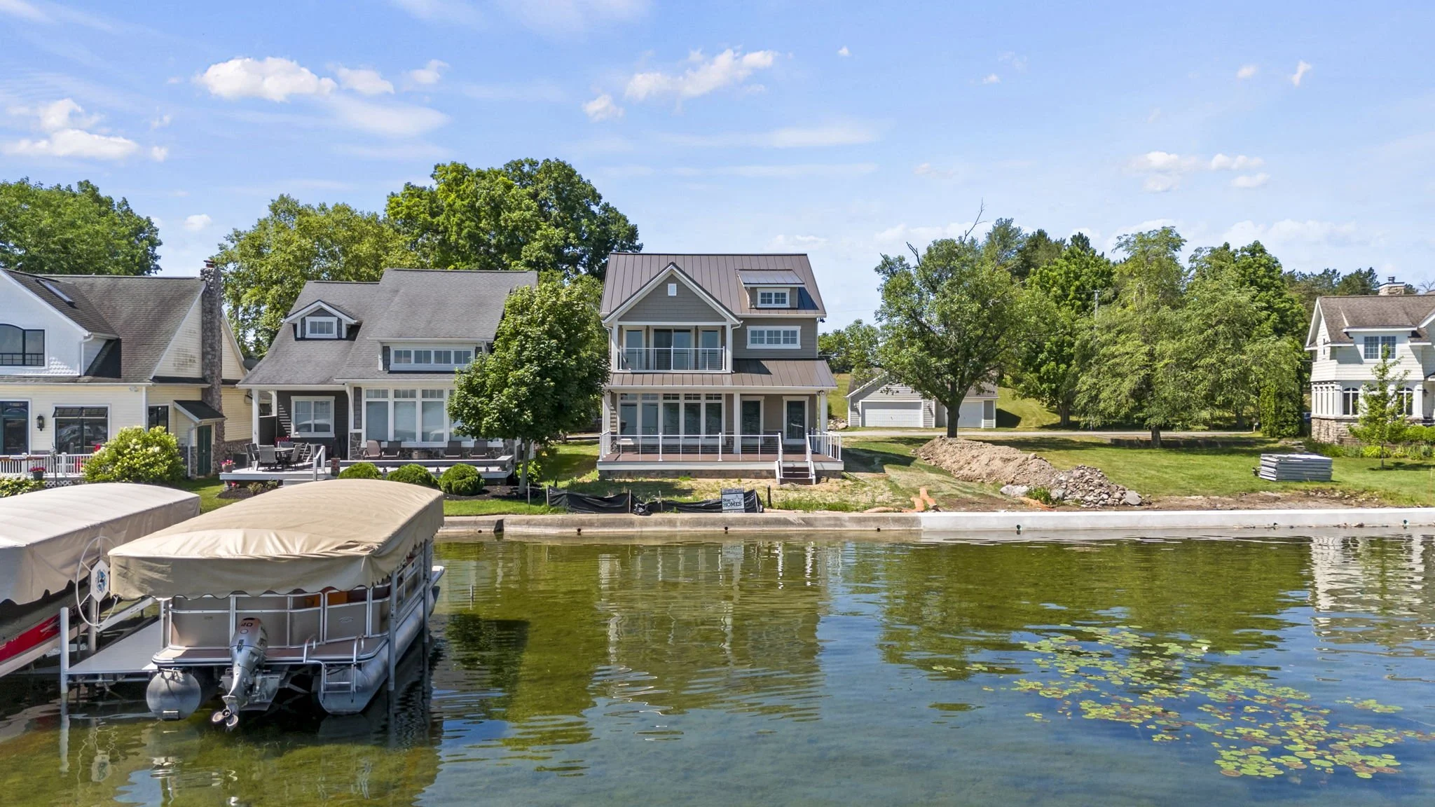 Residential houses along a waterfront, with boats docked in the water, green trees, and a partly cloudy sky.