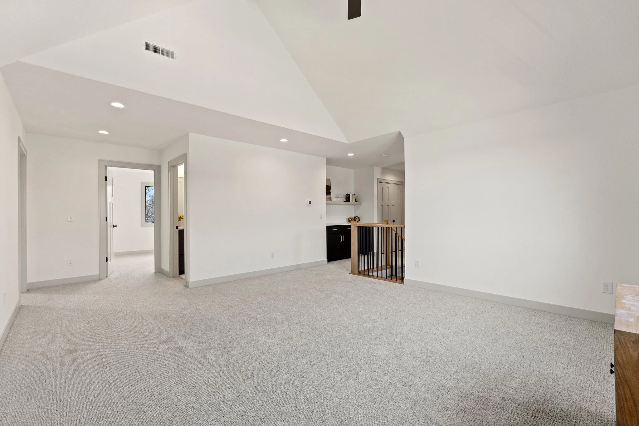 Open-concept living space with white walls, beige carpet, and recessed lighting. There are doorways leading to other rooms and a staircase with wooden handrails.