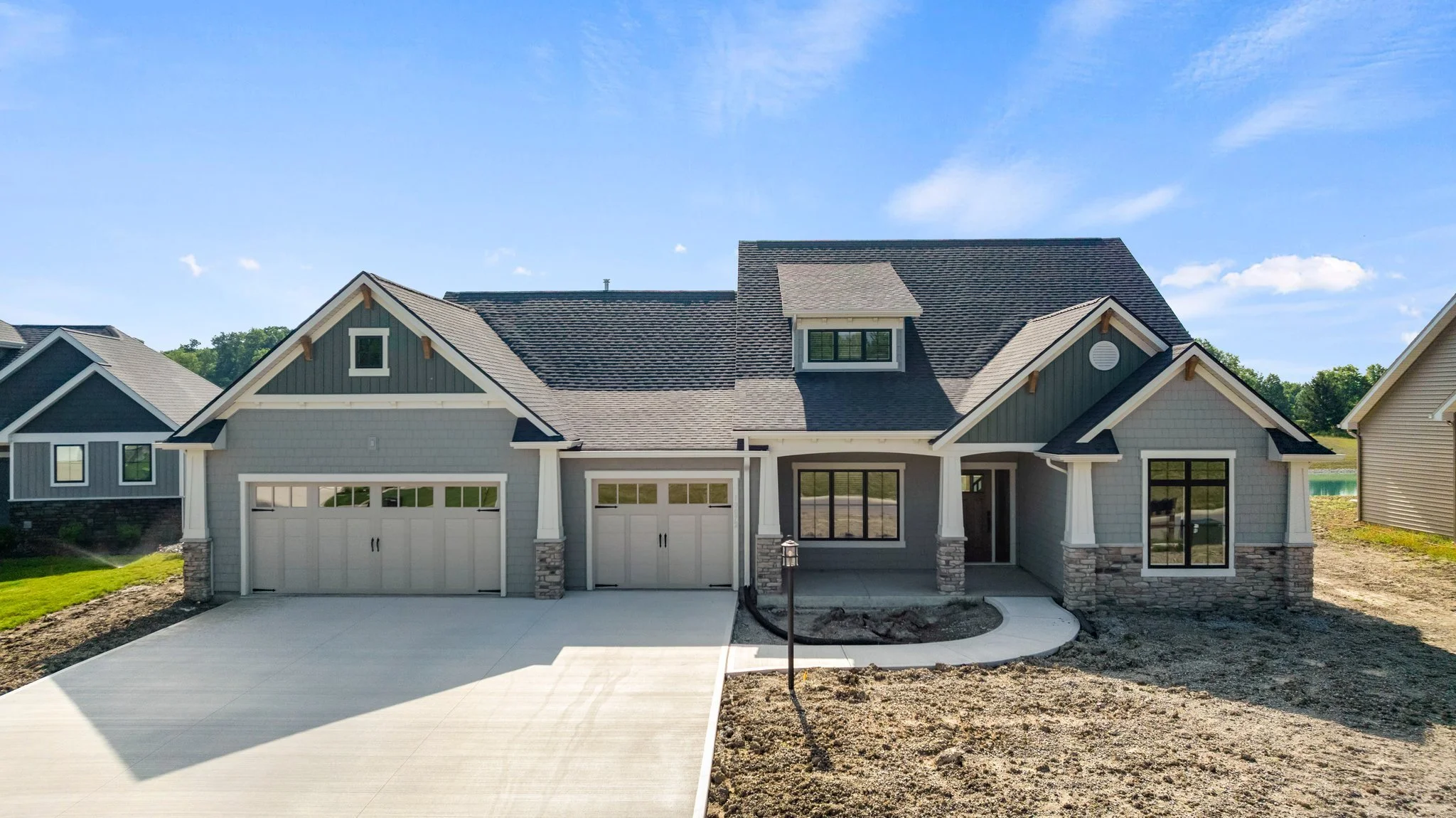 Newly constructed house with gray siding, stone accents, and a dark shingled roof, featuring a three-car garage, a covered front porch, and large windows, set against a clear blue sky.