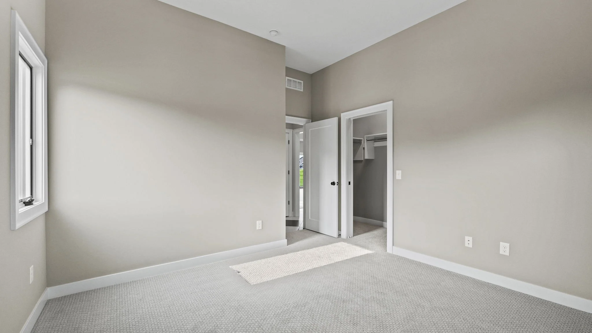 Empty room with gray walls, beige carpet, a window on the left, an open closet with white doors on the right, and a doorway leading to another room.