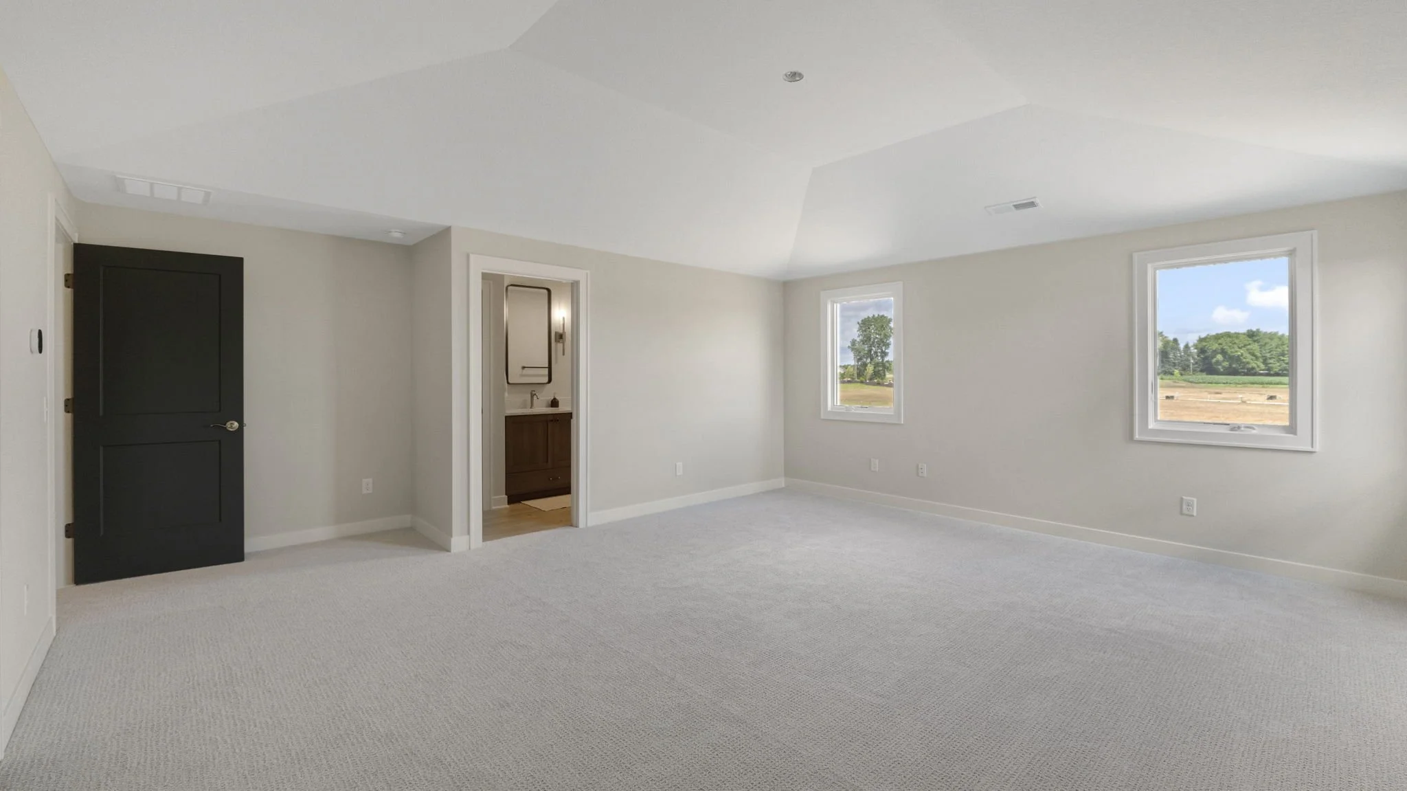Empty room with white walls and carpeted floor, two windows showing a grassy landscape, and an en-suite bathroom with a wooden vanity visible.