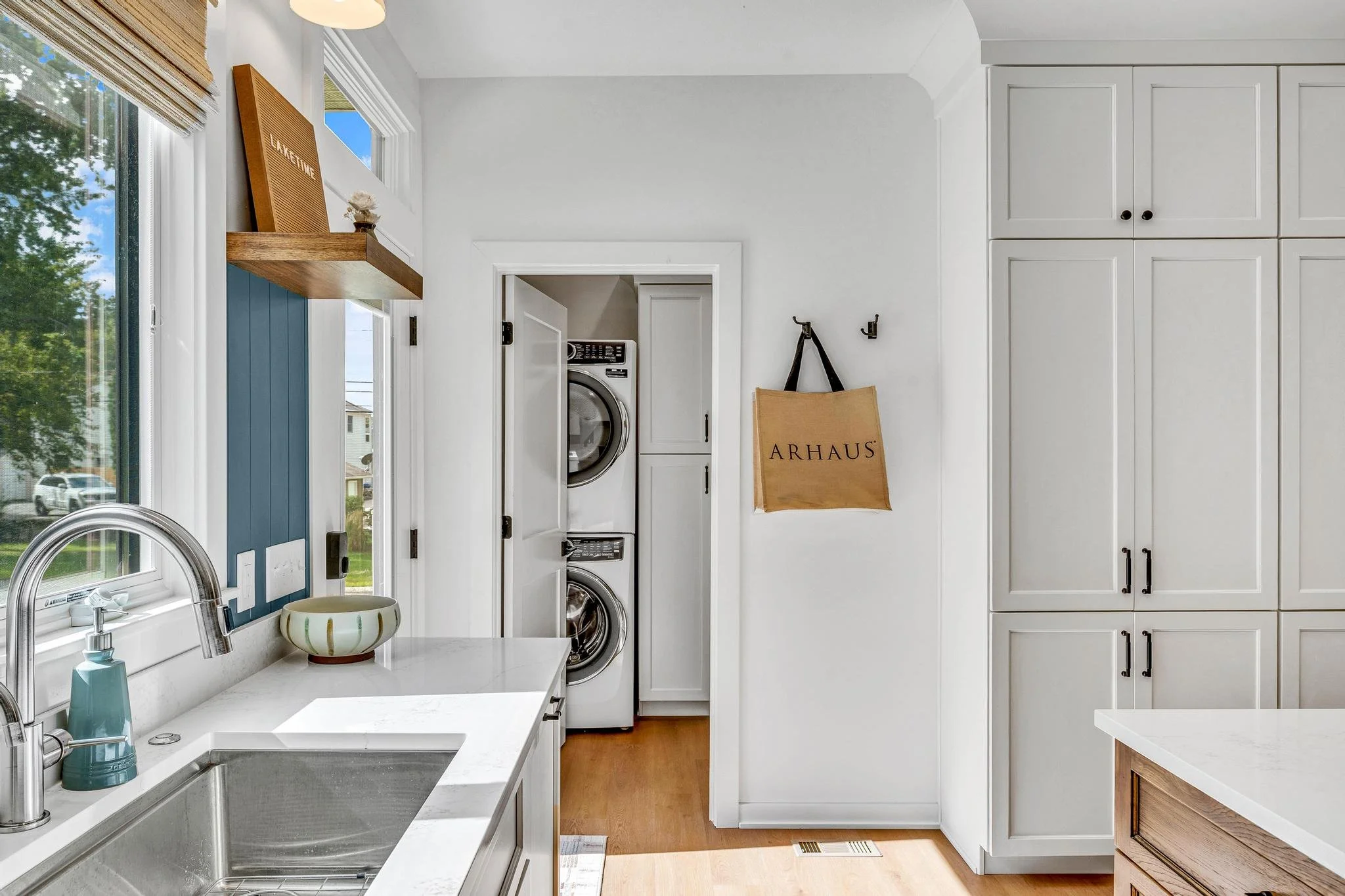 Kitchen with white cabinets, a window with blinds, a stainless steel sink, and a stacked washer and dryer in a small laundry area. A bag labeled 'ARHAUS' hangs on the wall.