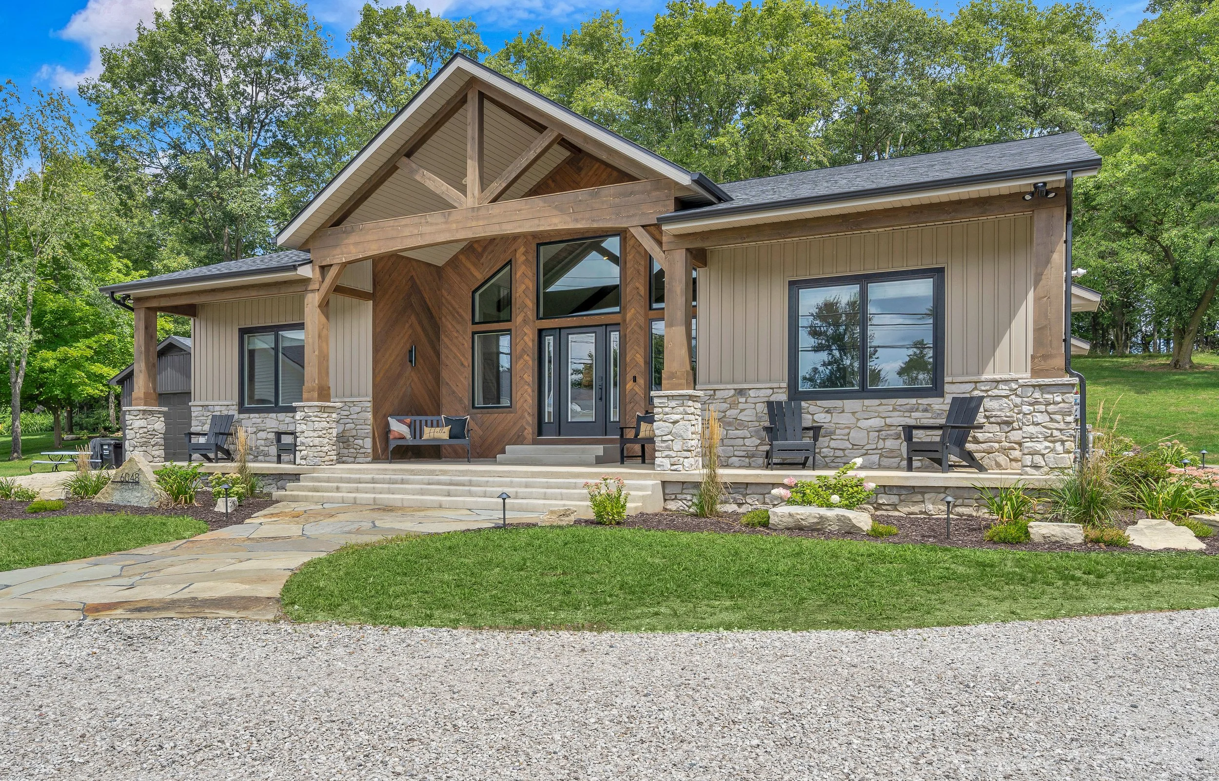 Front view of a modern house with a stone and wood facade, front steps, and a landscaped yard with shrubs and flowers, surrounded by trees.