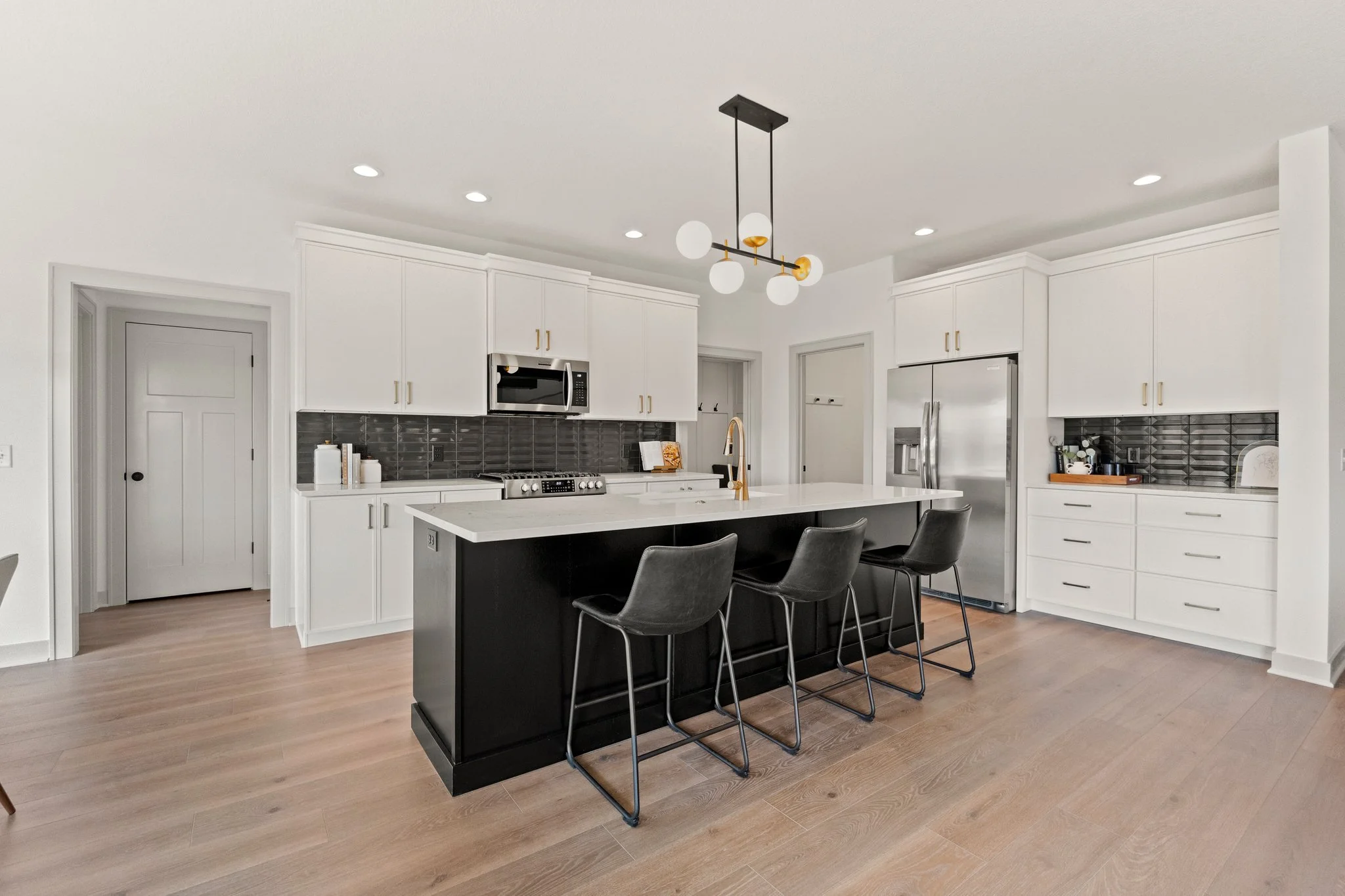 Modern kitchen with black island, white cabinets, gray tile backsplash, stainless steel appliances, and black barstools under a contemporary chandelier.