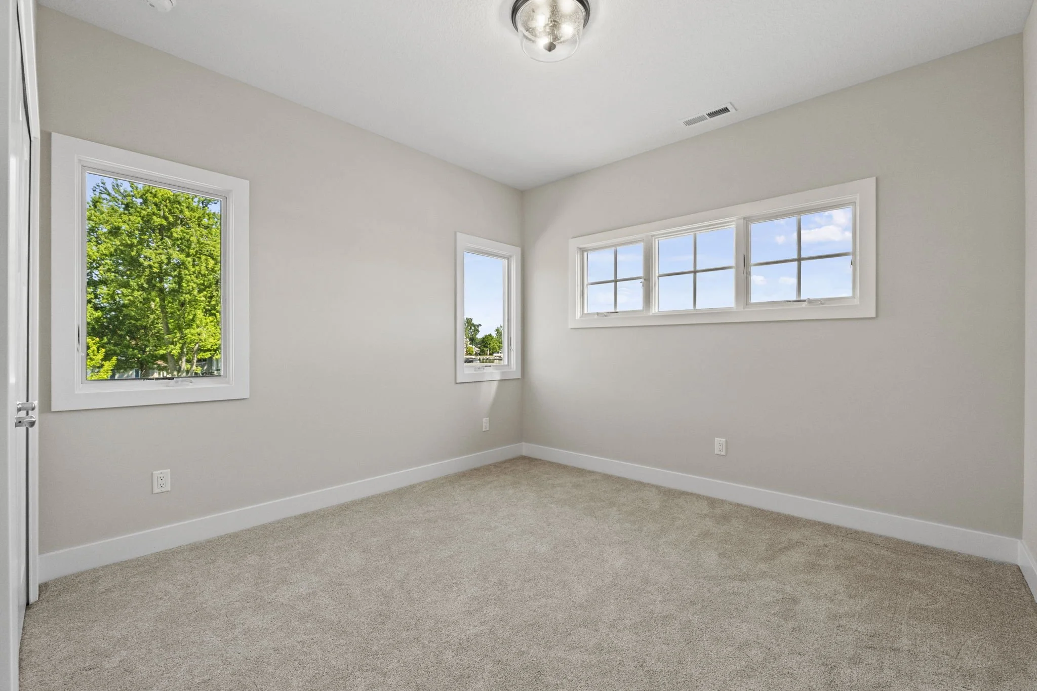 Empty room with beige carpet, white walls, three windows showing trees and sky, ceiling light, and electrical outlets.