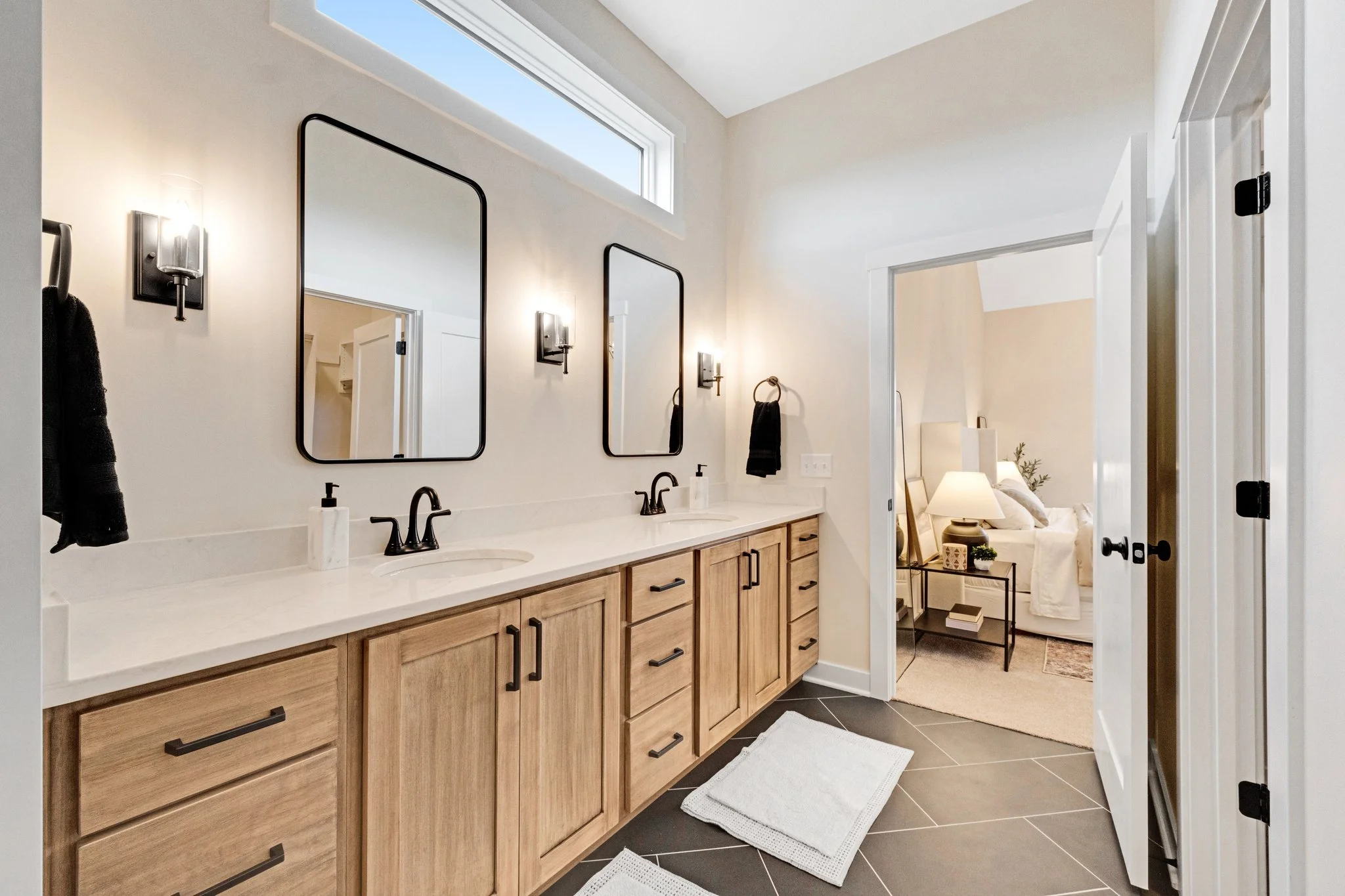Modern bathroom with dual sinks, wooden cabinets, black fixtures, and mirrors, leading to a bedroom with a lamp and bed.