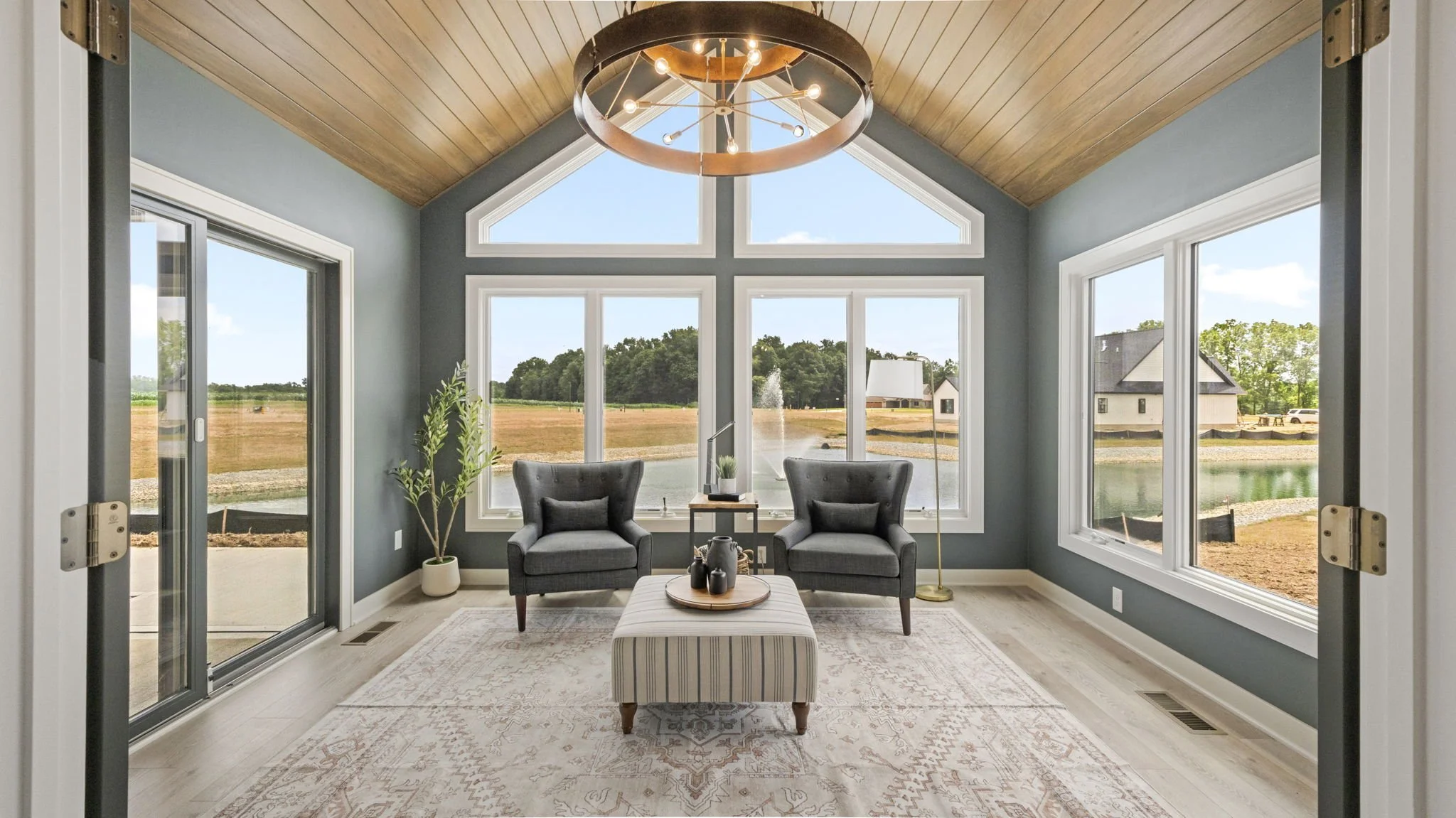 Living room with large windows overlooking a pond, two gray armchairs, a striped ottoman, a chandelier, and a wooden ceiling.