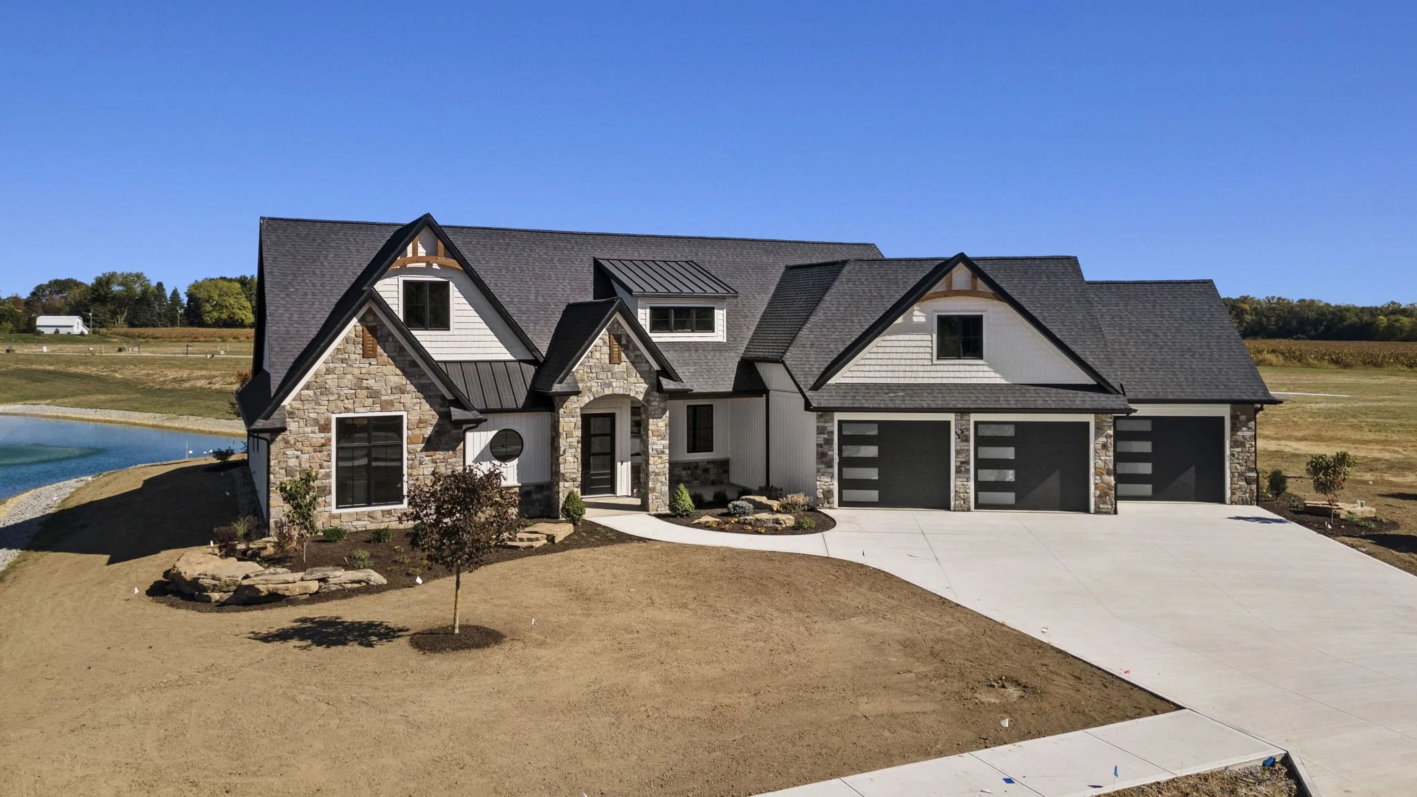 A modern house with stone and white siding exterior, black roof, three garage doors, and a concrete driveway, set against a clear blue sky with a pond to the left and open land in the background.