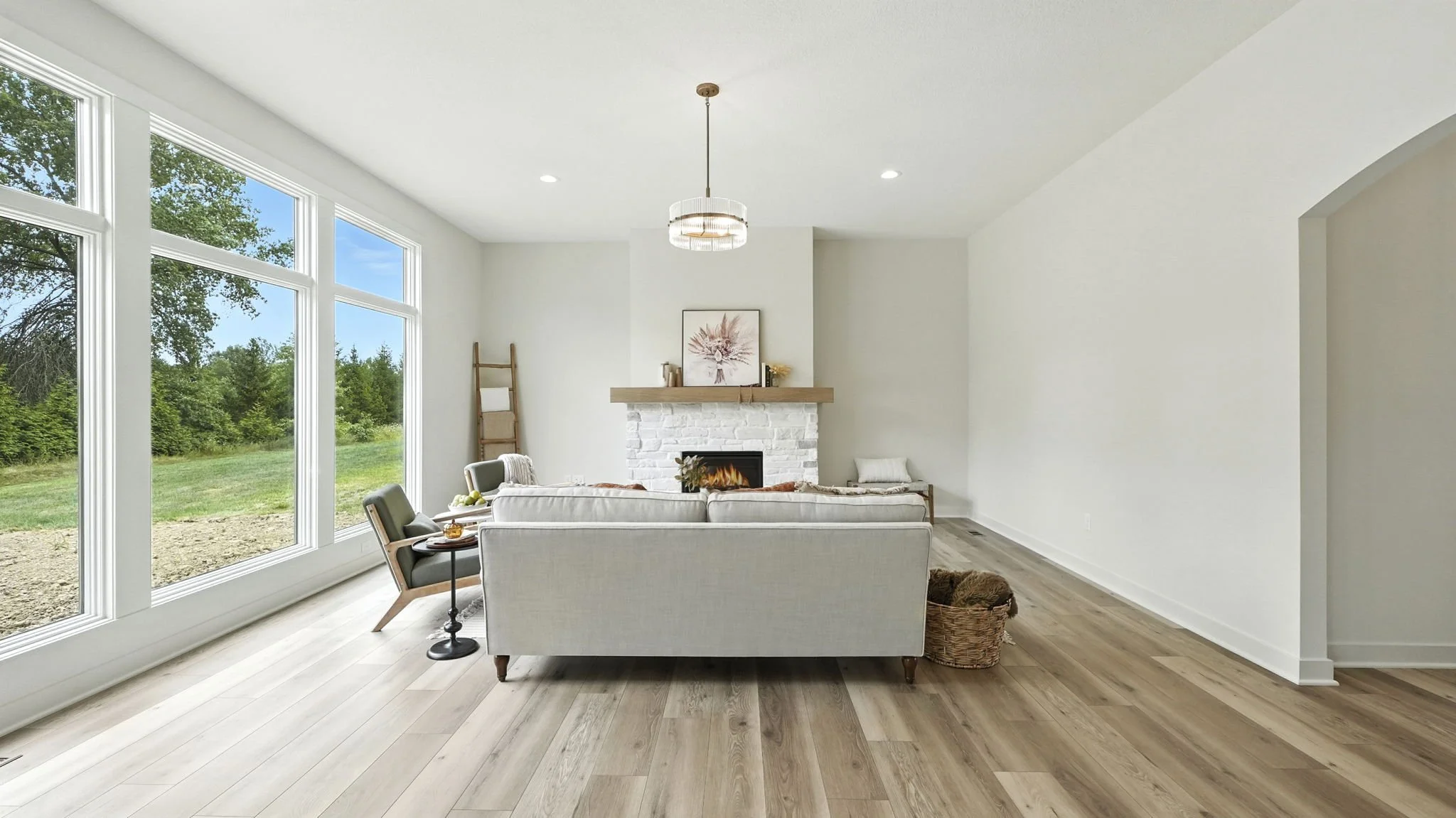Bright living room with large windows showing a green outdoor landscape, white walls, light wood flooring, a white couch, a chair, a basket with a blanket, a fireplace, and minimal decor.