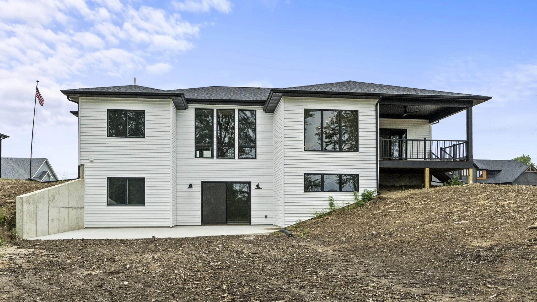 Newly constructed white two-story house with black accents, large windows, and a deck on the upper level, situated on a sloped dirt lot under a partly cloudy sky.