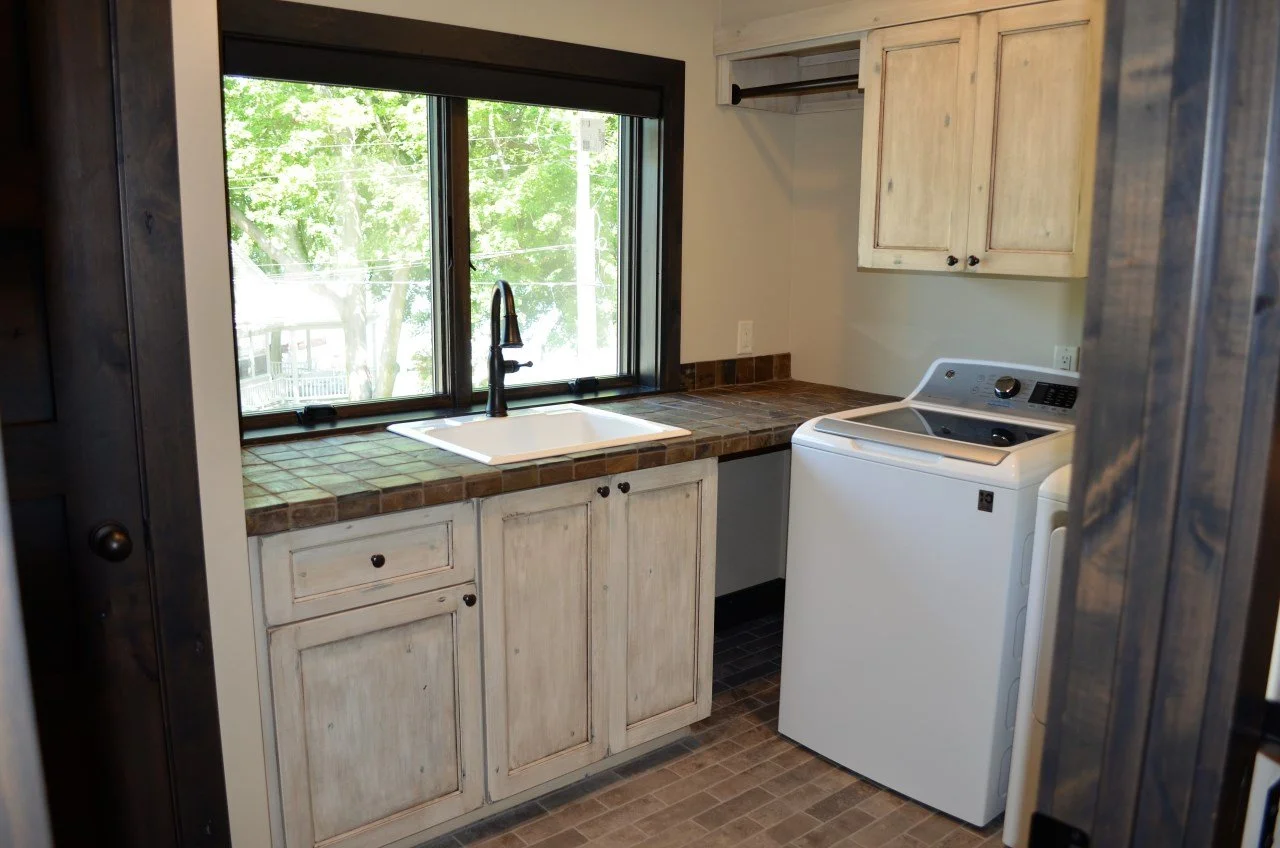 Laundry room with a window, a white washing machine, beige cabinets, a tiled countertop, a sink under the window, and a window with a view of green trees outside.
