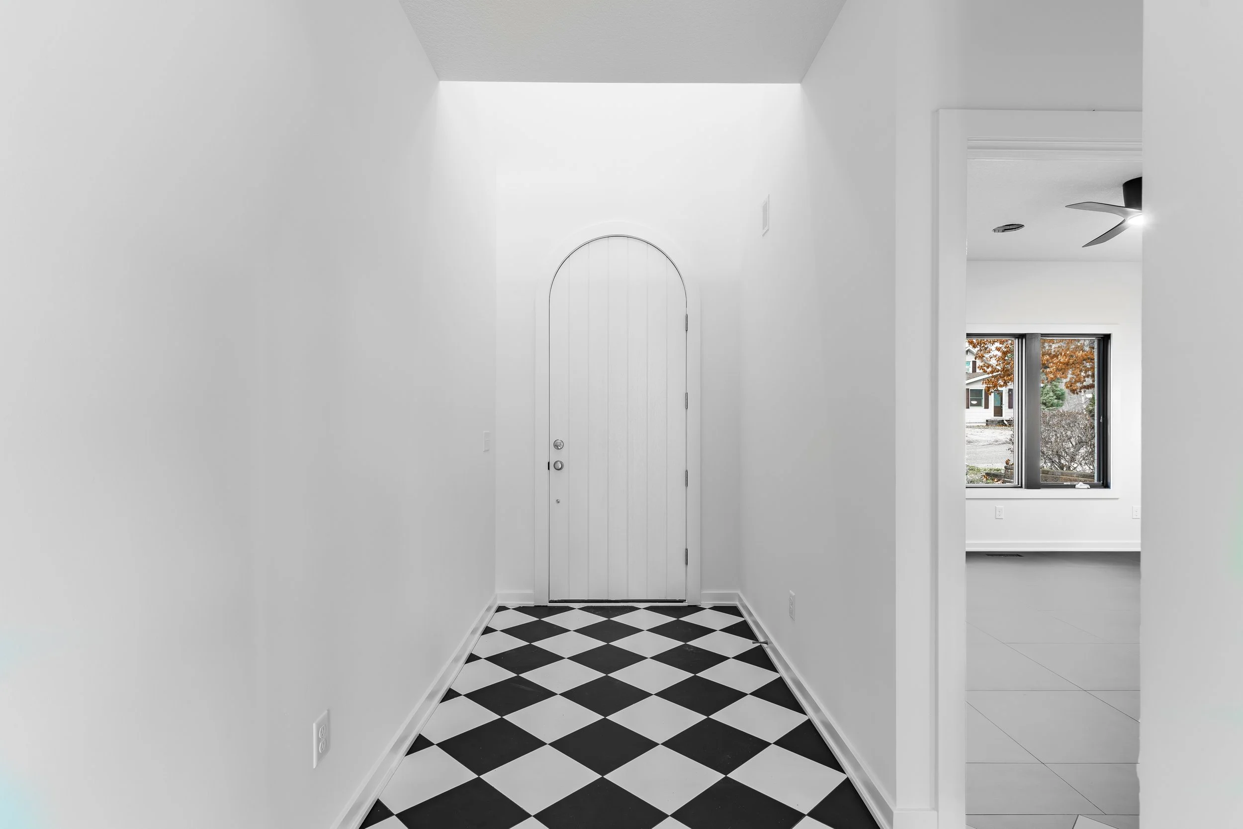 Entryway with a white arched door and black and white checkered floor, leading to a bright room with a window showing autumn trees outside.