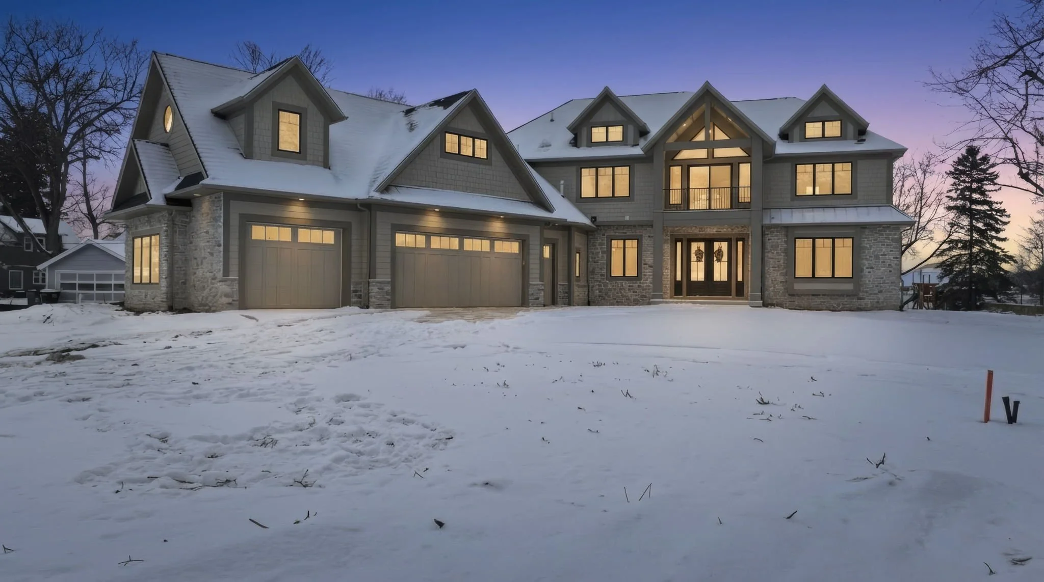 Large modern house with illuminated windows, snow-covered front yard, multiple garage doors, and a twilight sky.