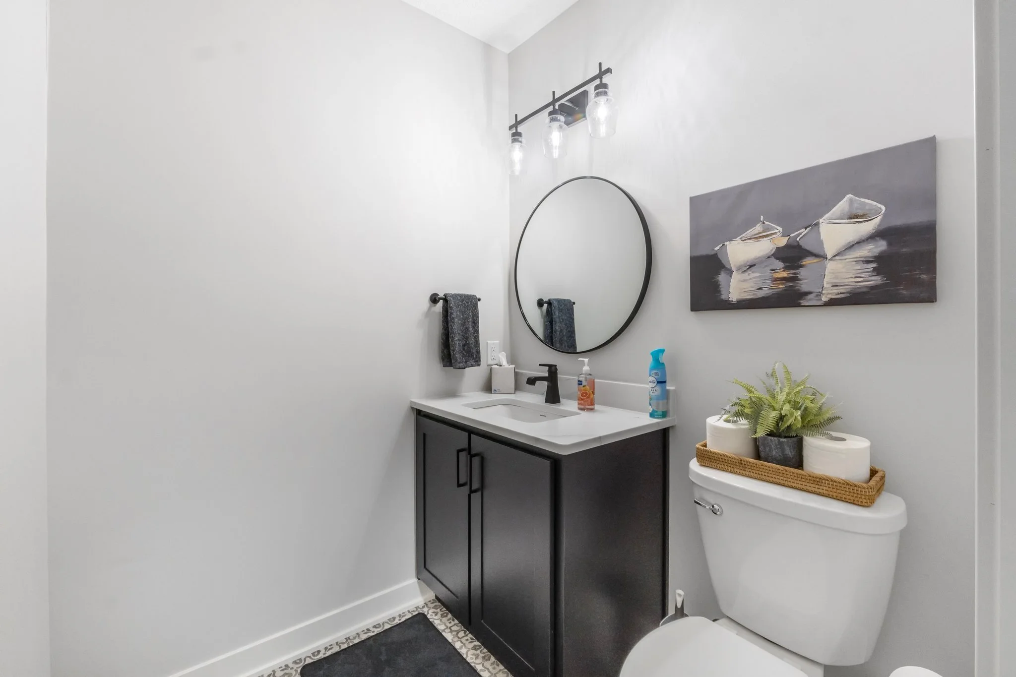 Modern bathroom with a black vanity, round mirror, and wall-mounted light fixture, featuring a toilet with a tray holding plants and toilet paper, and a wall art of two boats on the water.