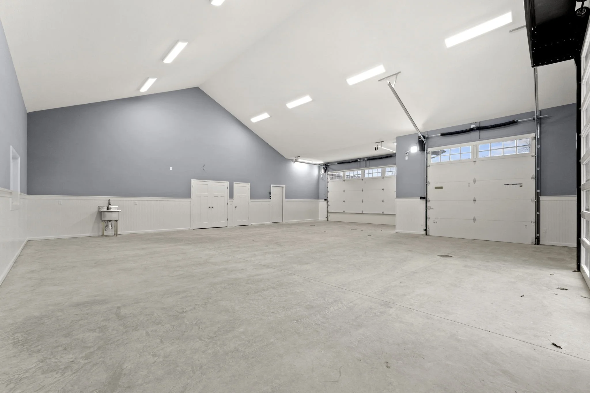 Empty garage with double white doors, concrete floor, gray upper walls, and white lower wall paneling, featuring small window at the top of the garage doors and a small utility sink on the left wall.