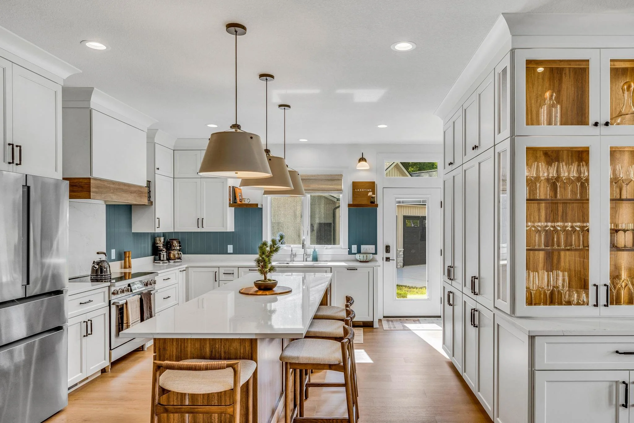 Modern kitchen with white cabinets, a large island with a white countertop, pendant lights, a French door, and glass-front cabinets displaying glassware.
