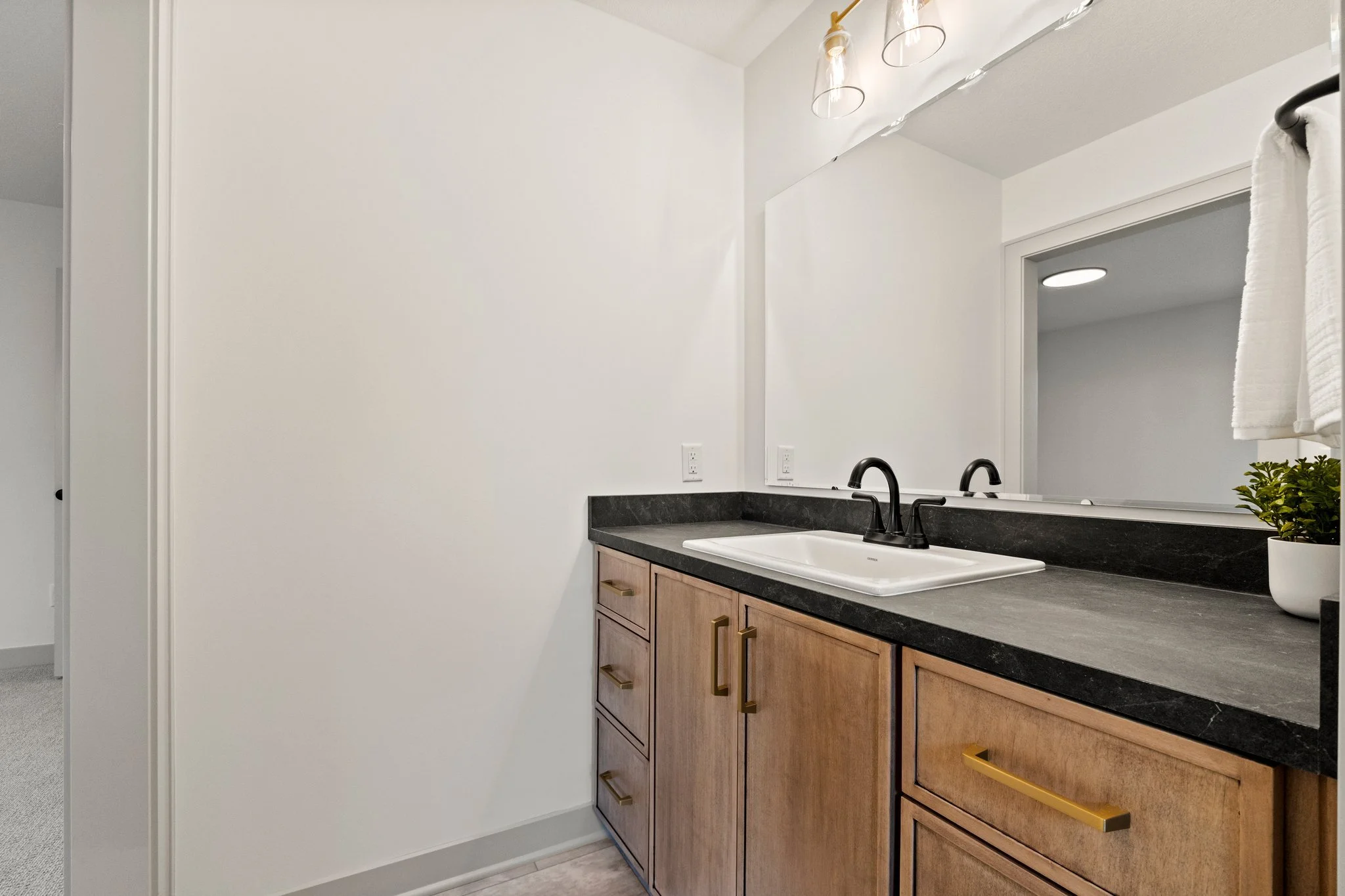 Modern bathroom with a black countertop, white sink, wooden cabinets, and a large mirror. There are white walls, a towel hanging, and some small plants on the counter. Overhead lights are visible in the reflection.
