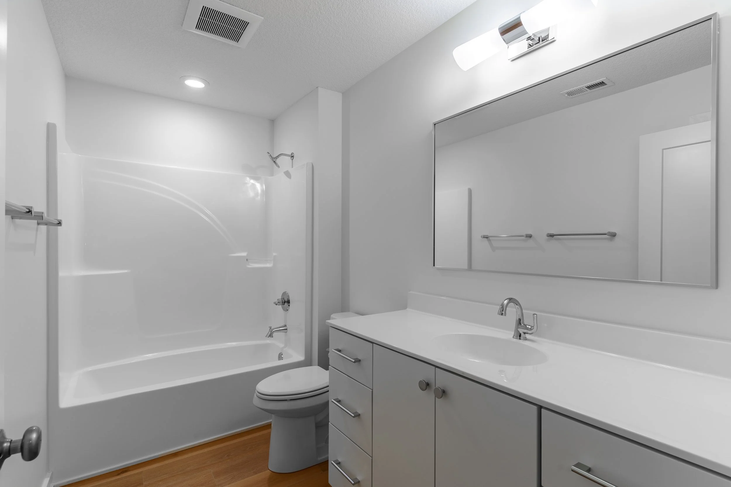 Modern bathroom with a white bathtub, toilet, and vanity with a large mirror and wall-mounted light, wooden flooring, and white walls.