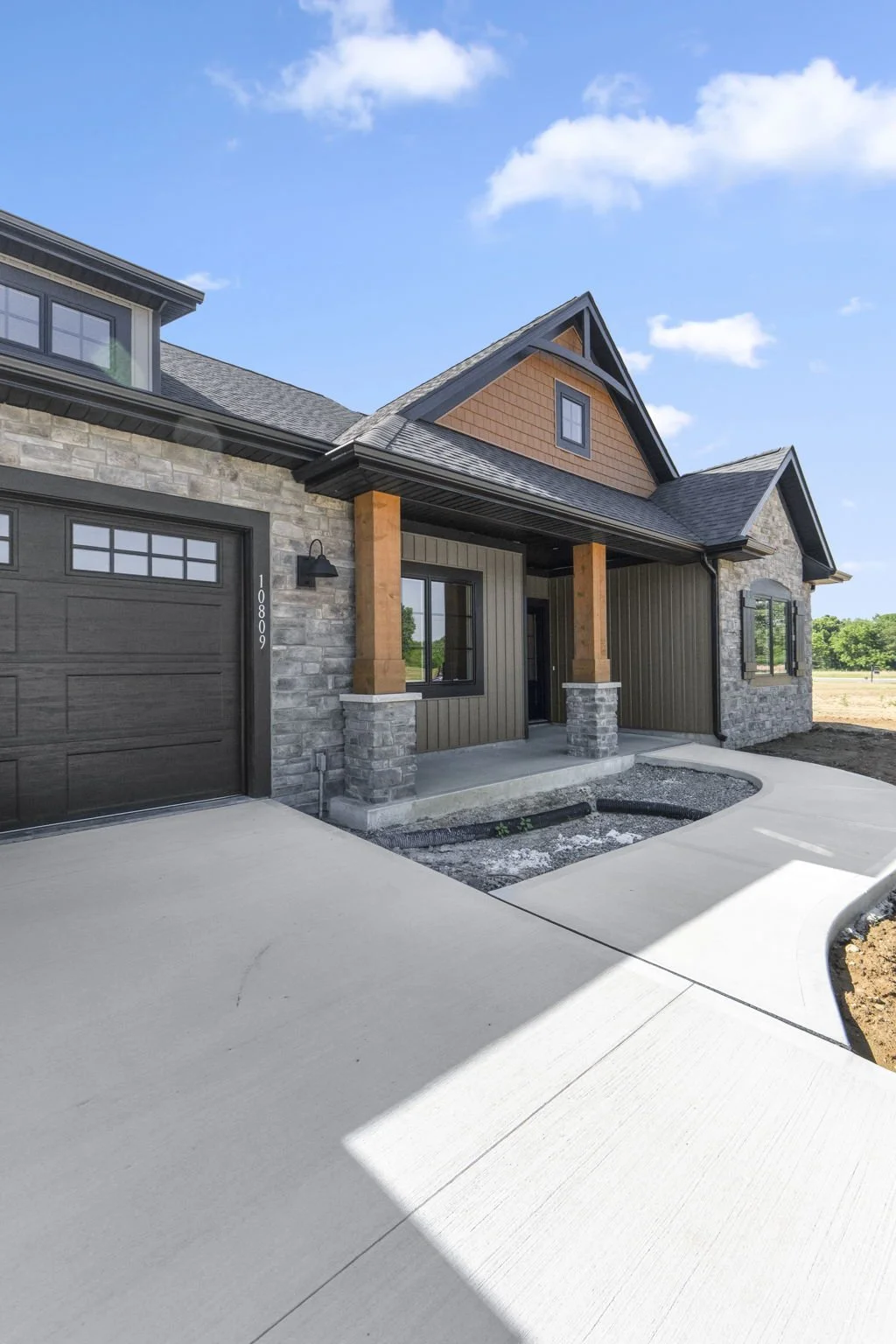 New modern house with graystone, dark siding, and wooden accents. Front porch with pillars, large windows, and a concrete driveway under a blue sky with clouds.