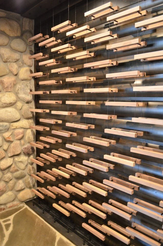 A decorative wooden shelf installation with multiple horizontal wooden slats attached to black vertical supports, mounted on a wall, with a stone wall on the left and a beige floor.