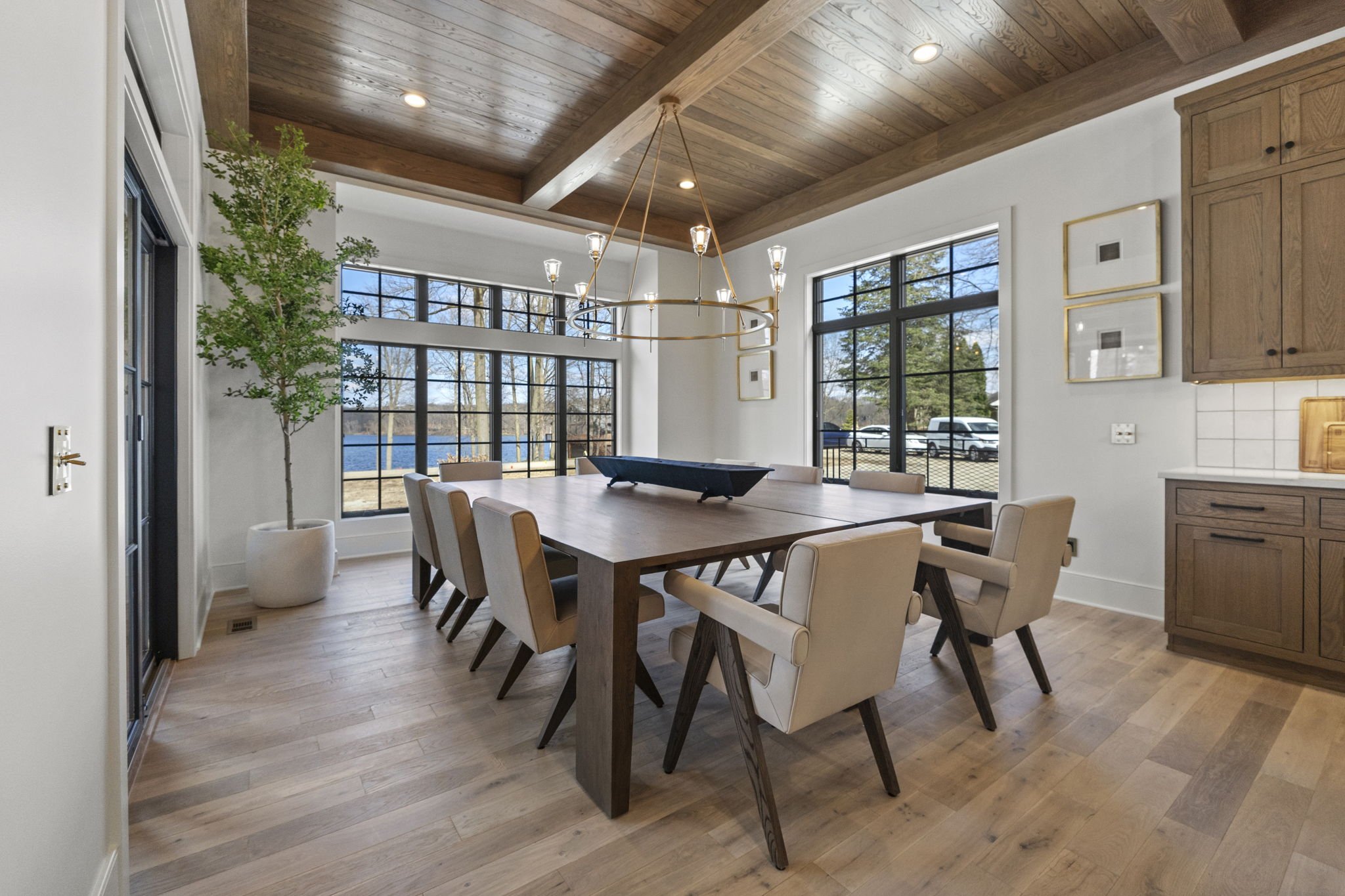 Dining room with wooden table and eight beige chairs, large windows, wooden ceiling, potted plant, chandelier, and view of outdoors.