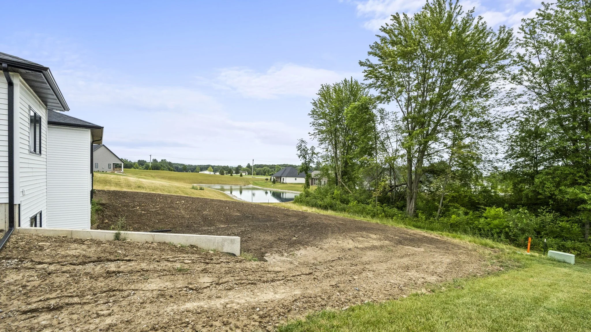 View of a backyard under construction with a patch of dirt, a white house on the left, green trees on the right, a lake, and houses in the distance under a blue sky with some clouds.