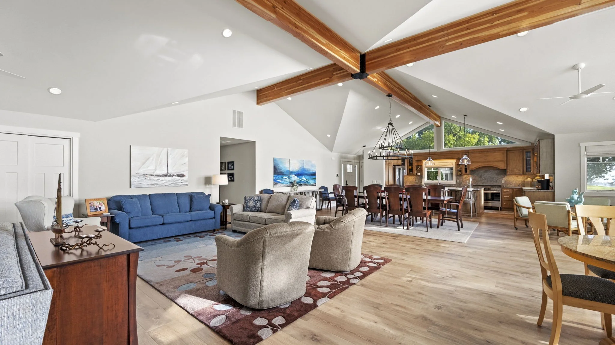 Open-concept living and dining room with wooden ceiling beams, white walls, hardwood flooring, and large windows. The living area has a blue sofa, beige and gray armchairs, and a patterned rug. The dining area features a long wooden table with seatin