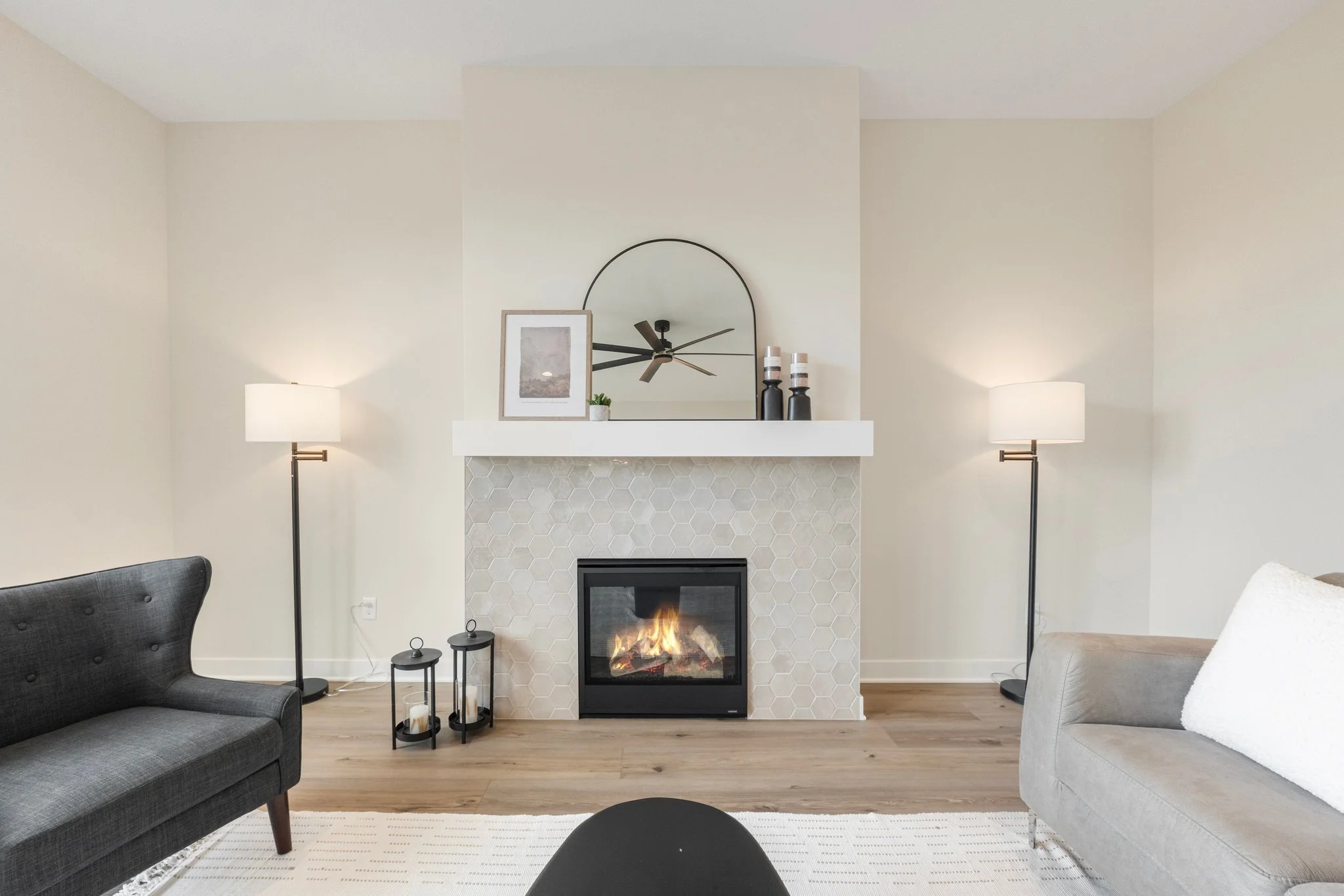 Living room with a fireplace, two floor lamps, a mirror above the fireplace, and two sofas, one black and one light gray, on a wooden floor.