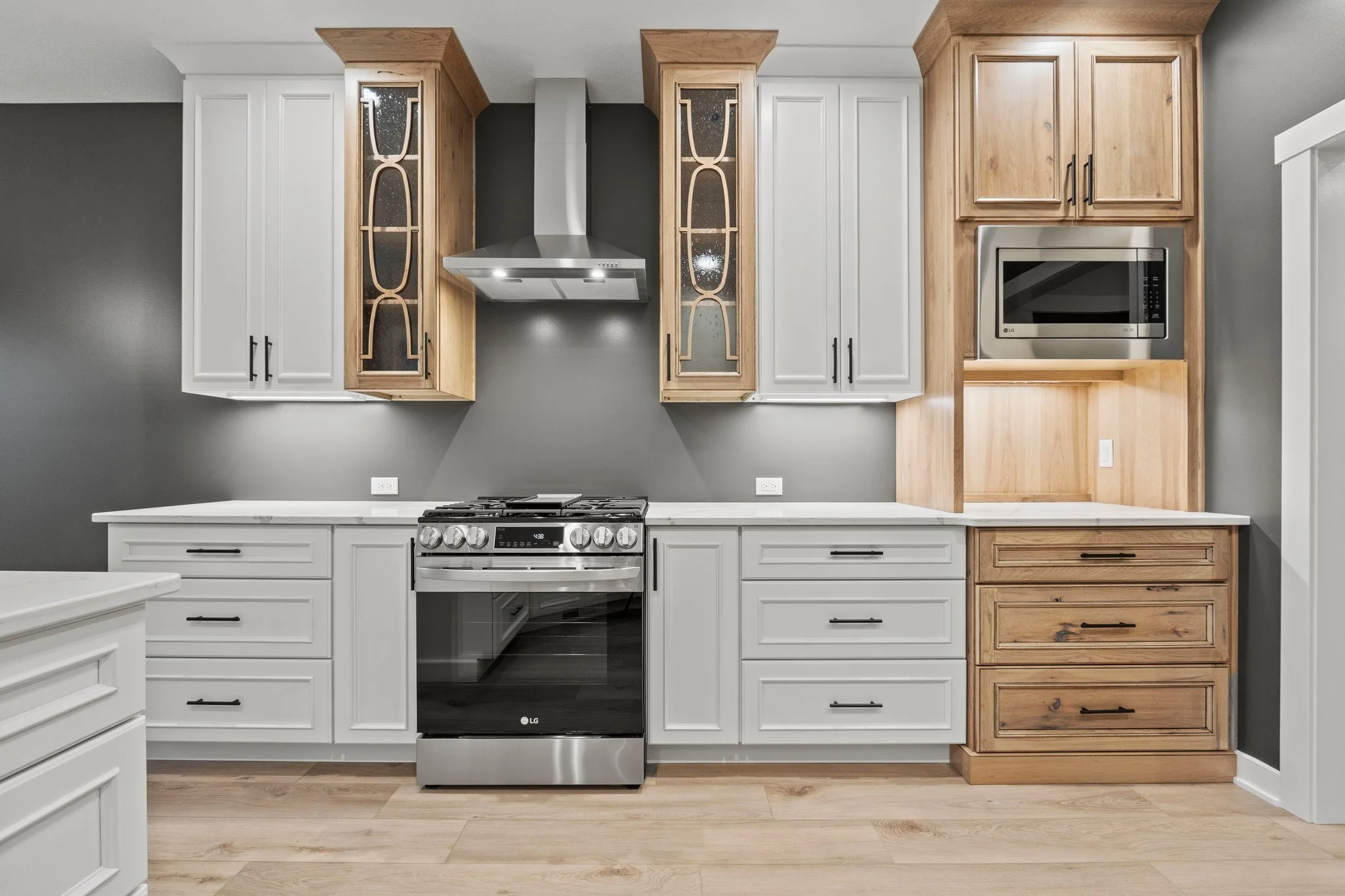 Modern kitchen with white and natural wood cabinetry, stainless steel stove and microwave, gray wall, light wood flooring, and decorative glass-front cabinets.