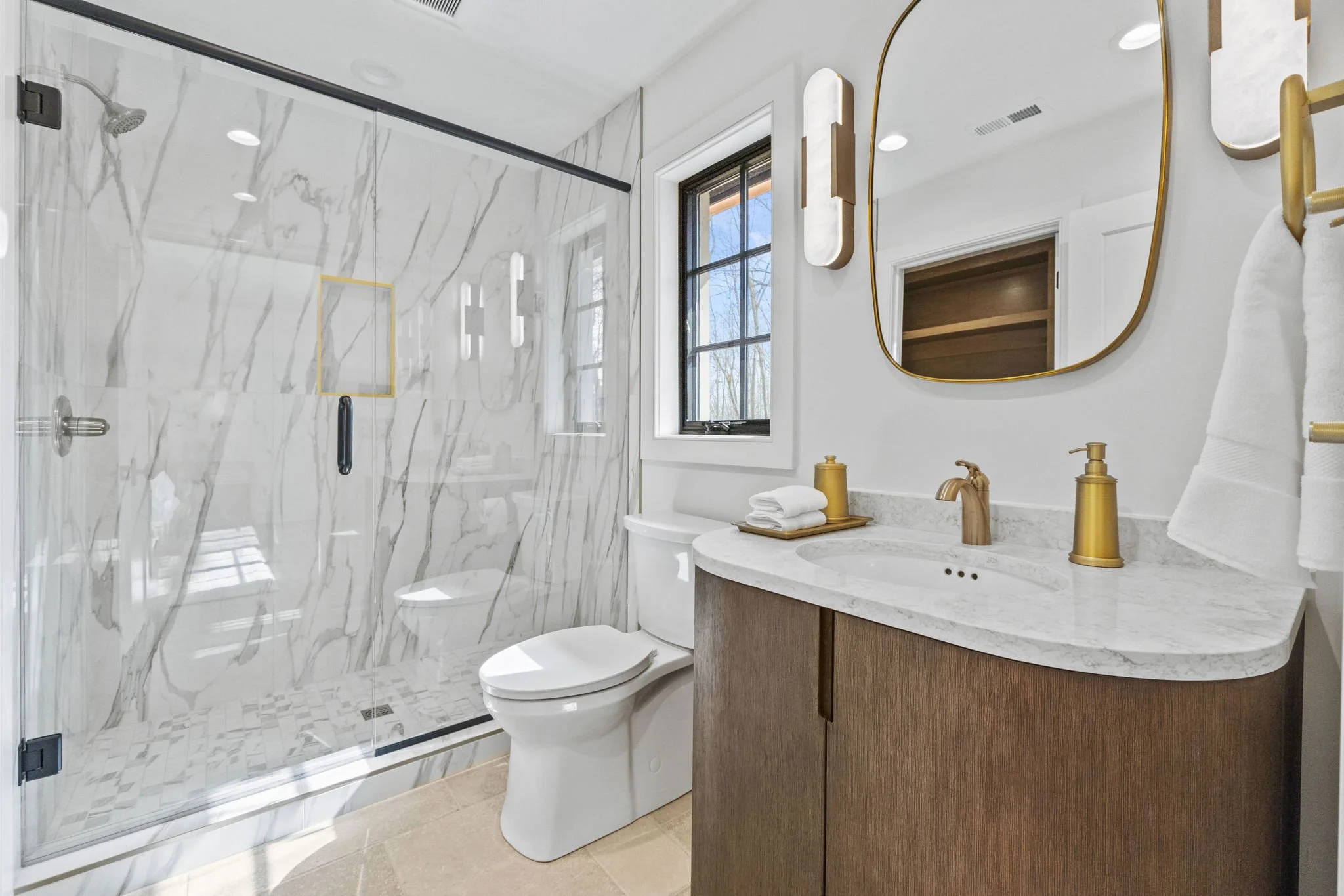Modern bathroom featuring a glass-enclosed walk-in shower with marble tile, a white toilet, a wooden vanity with a marble countertop, a gold faucet, matching gold soap dispenser and towel holder, a rectangular mirror with a gold frame, a window with 