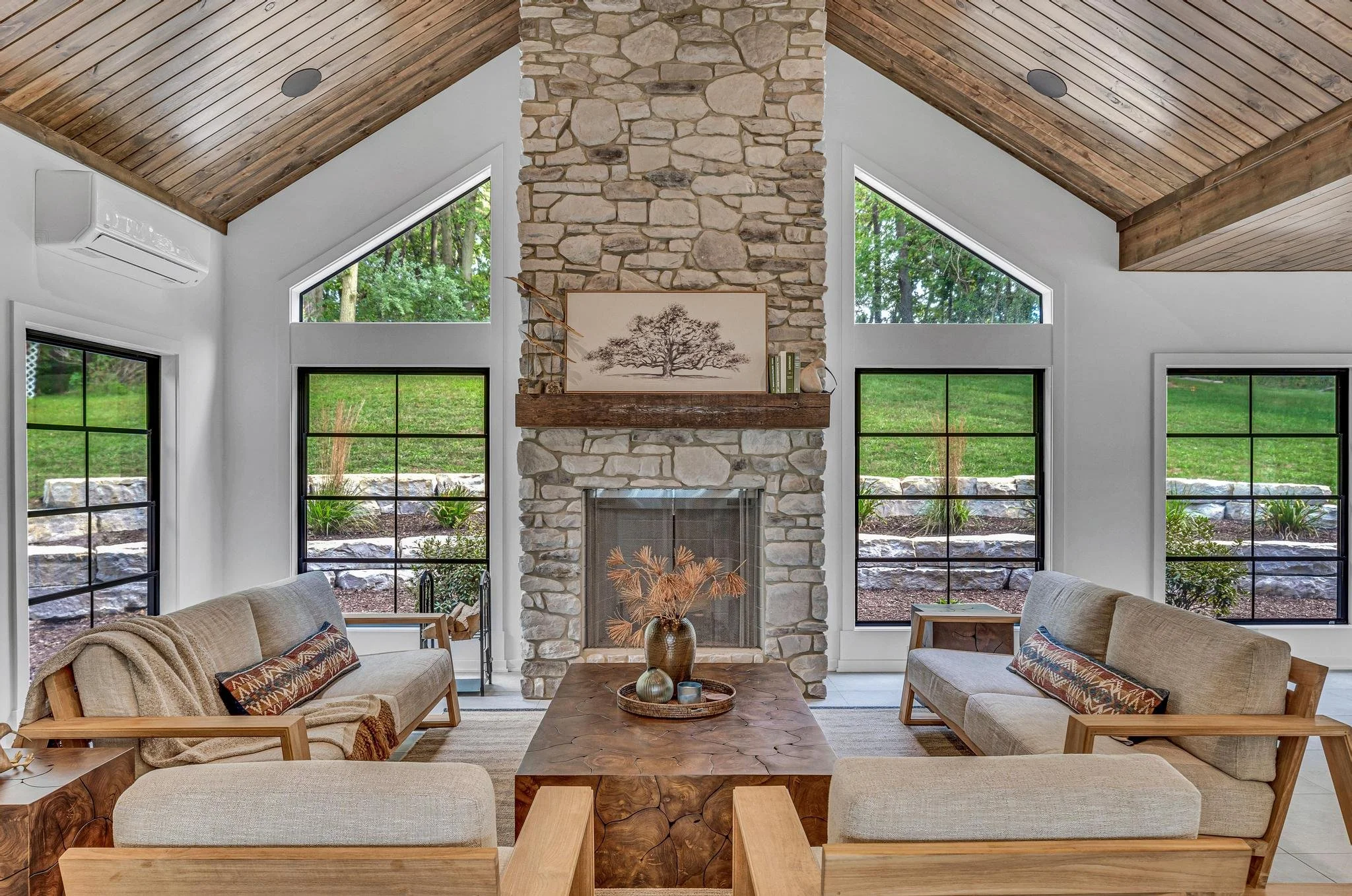 Living room with a stone fireplace, wooden ceiling, and large windows showing greenery outside. Four beige sofas with wooden armrests surround a wooden coffee table, decorated with vases and dried plants.