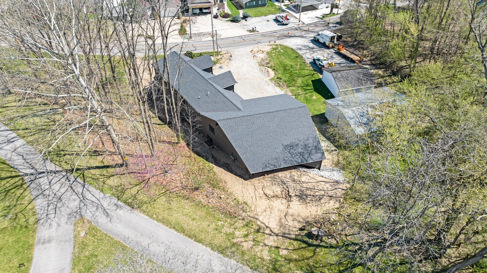 Aerial view of a residential area showing a black house with a gray roof under construction, surrounded by leafless trees and a landscaped yard, with construction vehicles and equipment nearby.