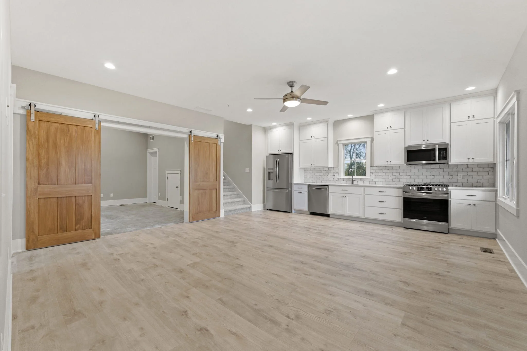 Open kitchen and living room with white cabinets, stainless steel appliances, light wood flooring, and a ceiling fan with lighting.