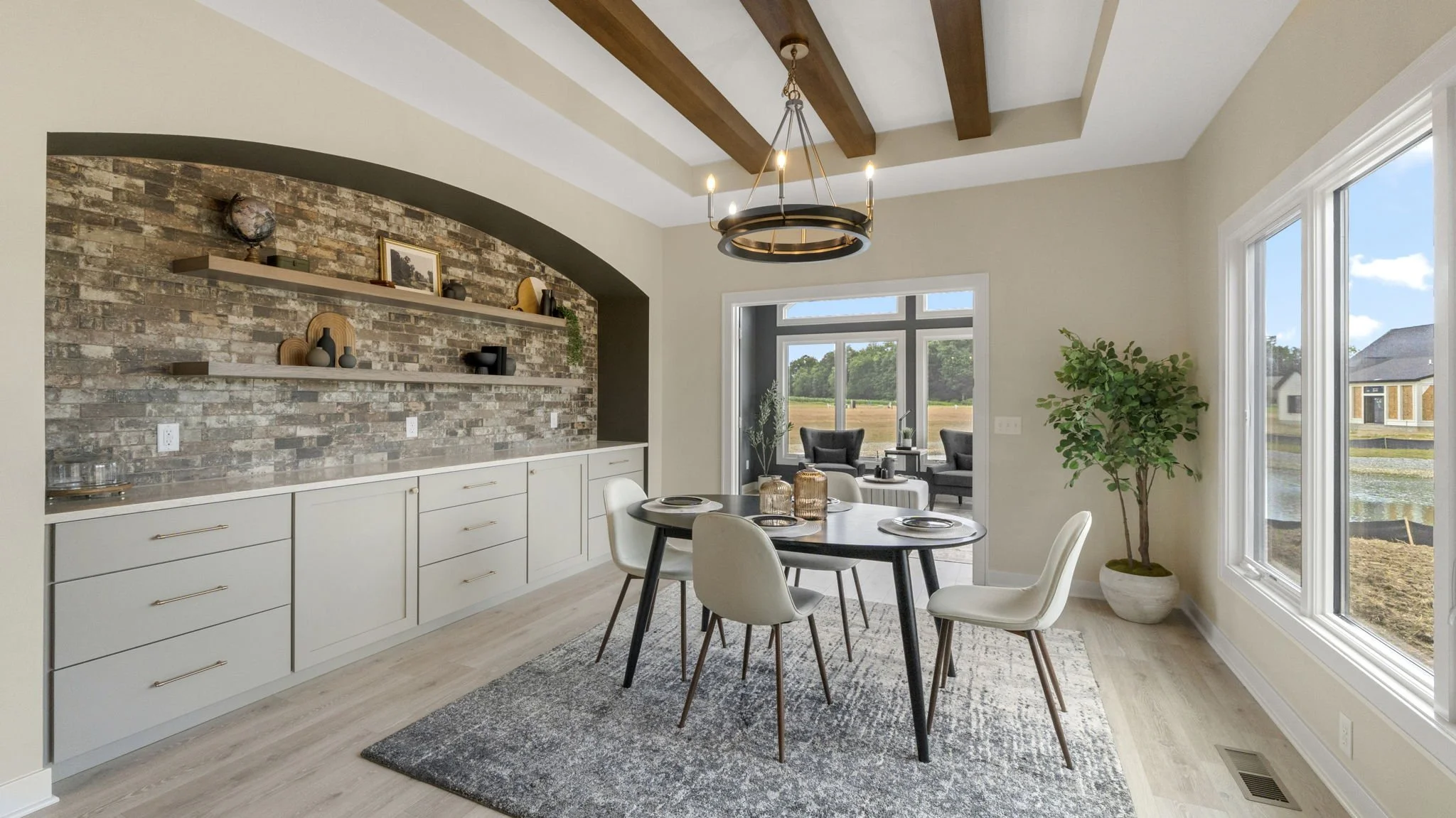 Dining area with a round black table, six white chairs, a hanging chandelier, a brick accent wall with floating shelves holding decorative objects, a large window showing outdoor scenery, and a potted plant in the corner.