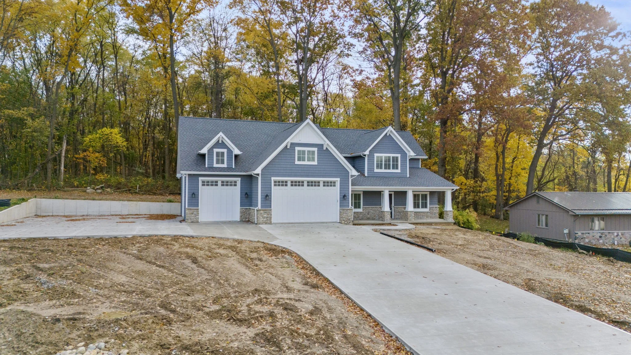 New blue two-story house with white trim, stone accents, and a two-car garage, surrounded by fallen leaves and woods.