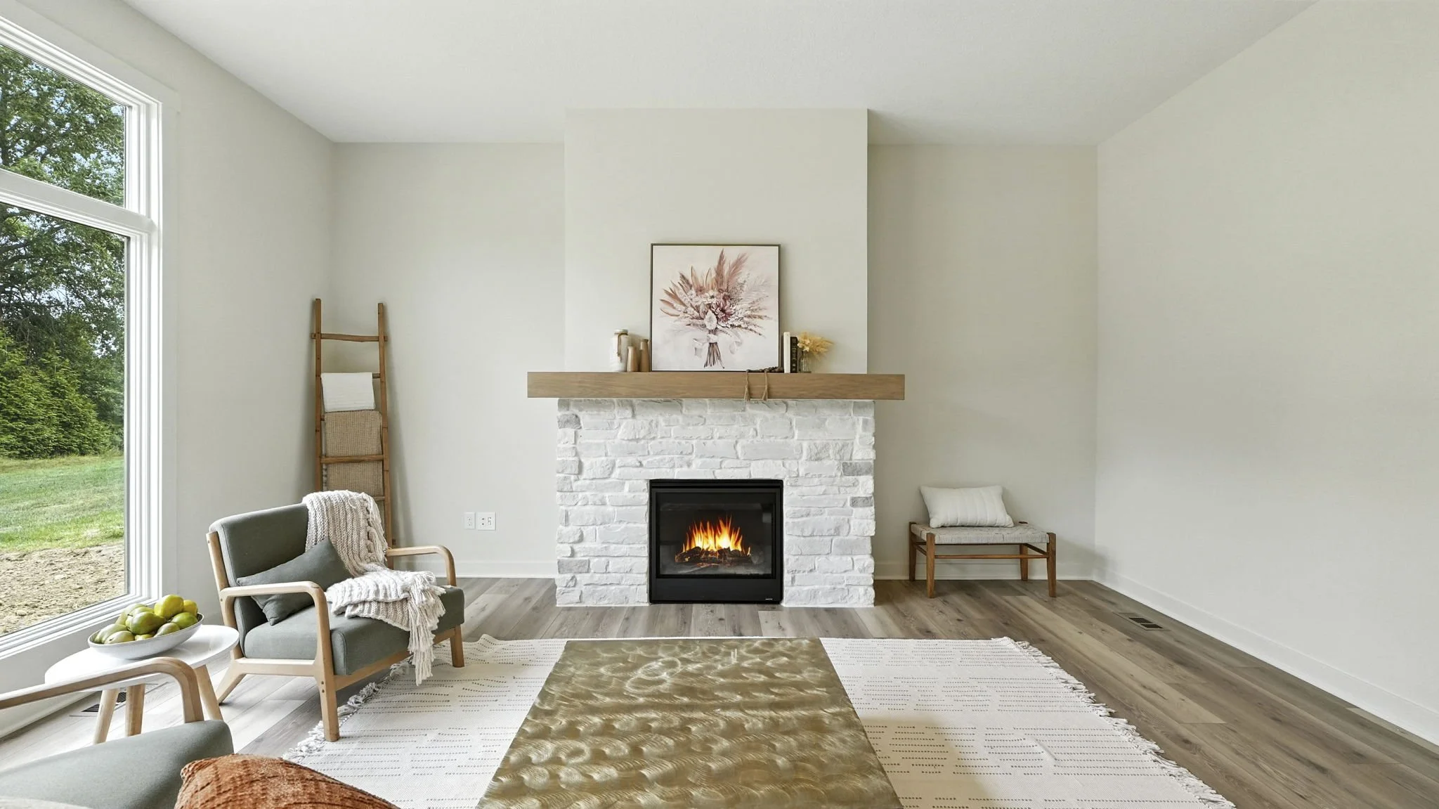A modern living room with a white brick fireplace, a wooden mantel, and a fire burning inside. To the left, large windows showing a green outdoor landscape. The room features minimalist furniture, including a gray armchair with a blanket, a wooden be