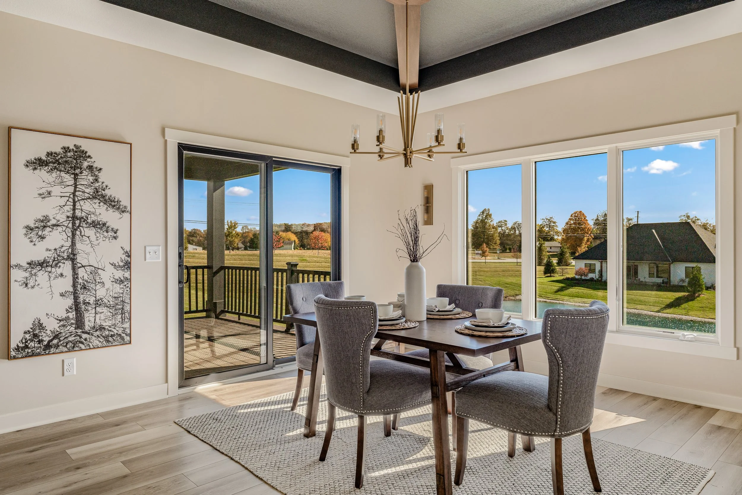 Dining room with a wooden table, six gray upholstered chairs, a white vase with branches, large windows showing an outdoor grass yard and houses, and a sliding glass door leading to a balcony.