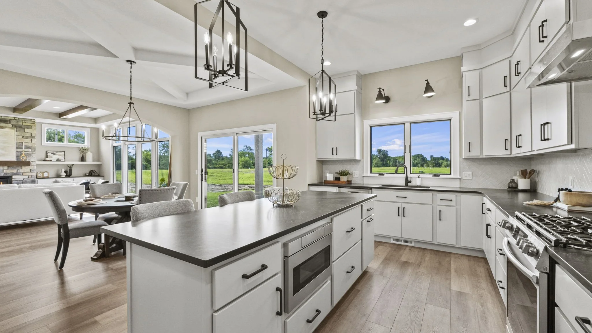 Bright kitchen with white cabinets, black hardware, black countertops, and a kitchen island with a microwave. Open layout with dining and living area, large windows and sliding door showing greenery outside. Modern light fixtures hanging from the cei
