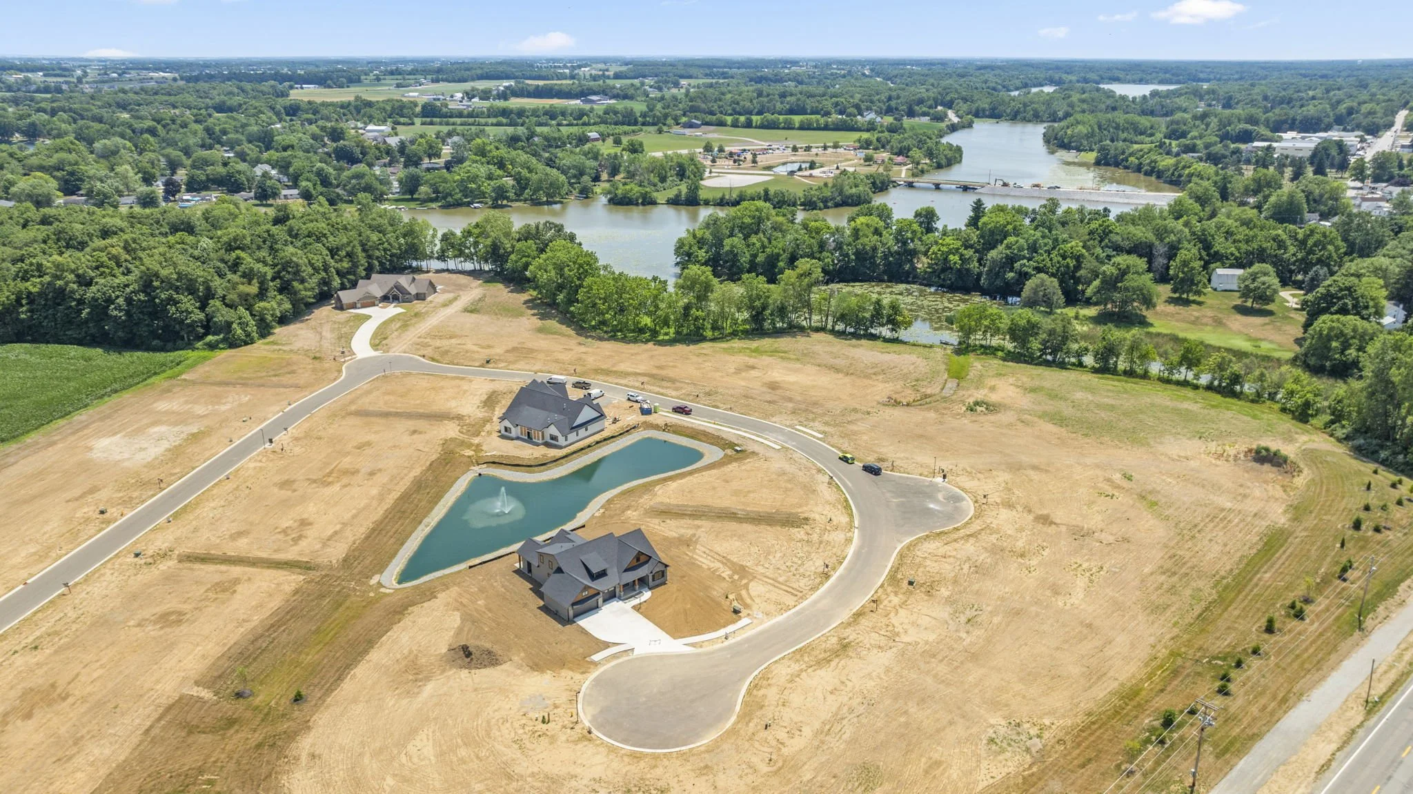 Aerial view of a residential area with a house, pond with fountain, and empty land, near a river and wooded areas.